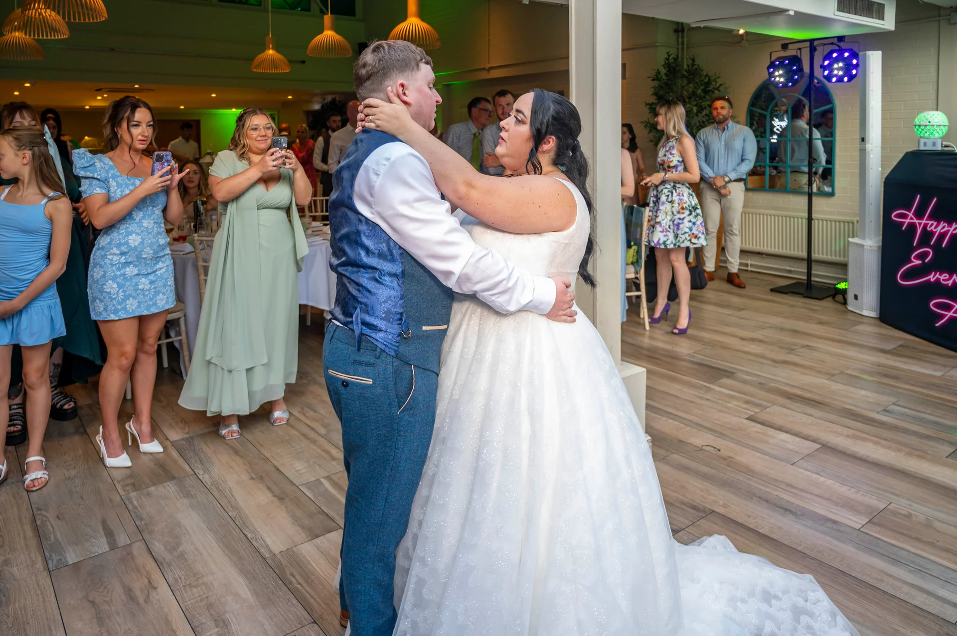 The newlywed couple share a tender embrace during their first dance while wedding guests watch and capture the moment on their phones.