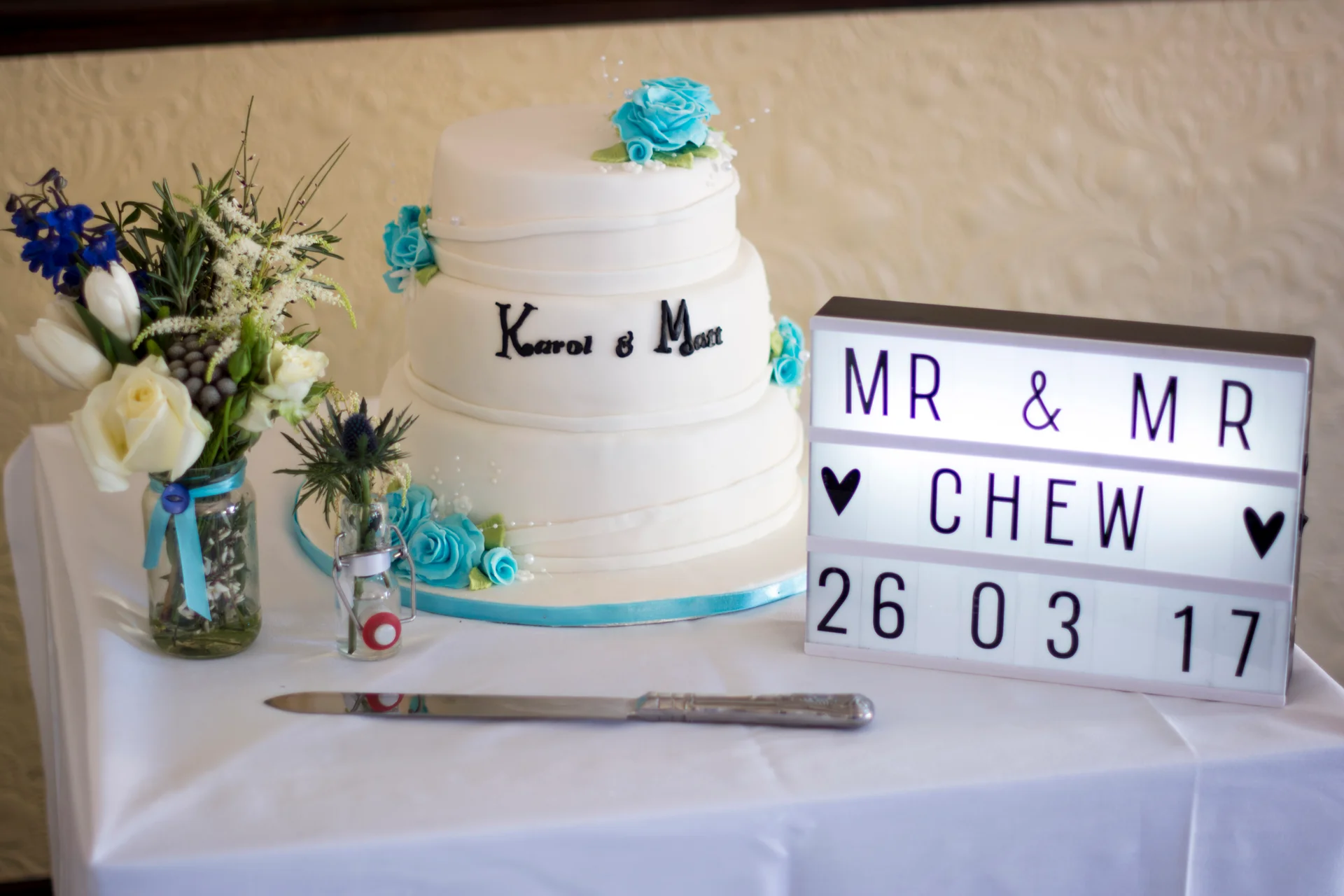 A three-tier white wedding cake decorated with blue sugar roses sits next to a glowing lightbox and floral arrangement on a white-clothed table.