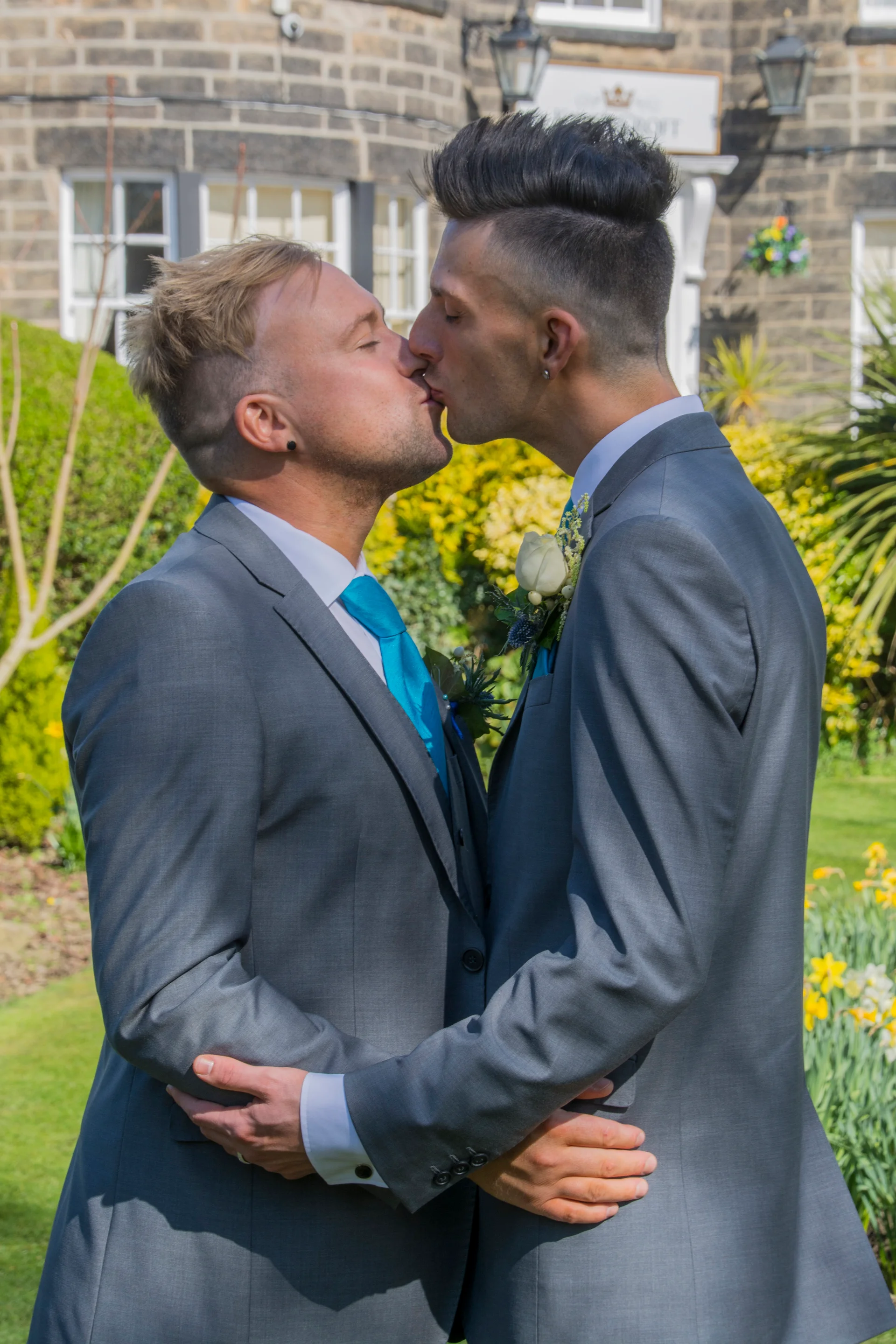 Two grooms in matching grey suits and blue ties share a romantic first kiss outdoors in a sunlit garden.