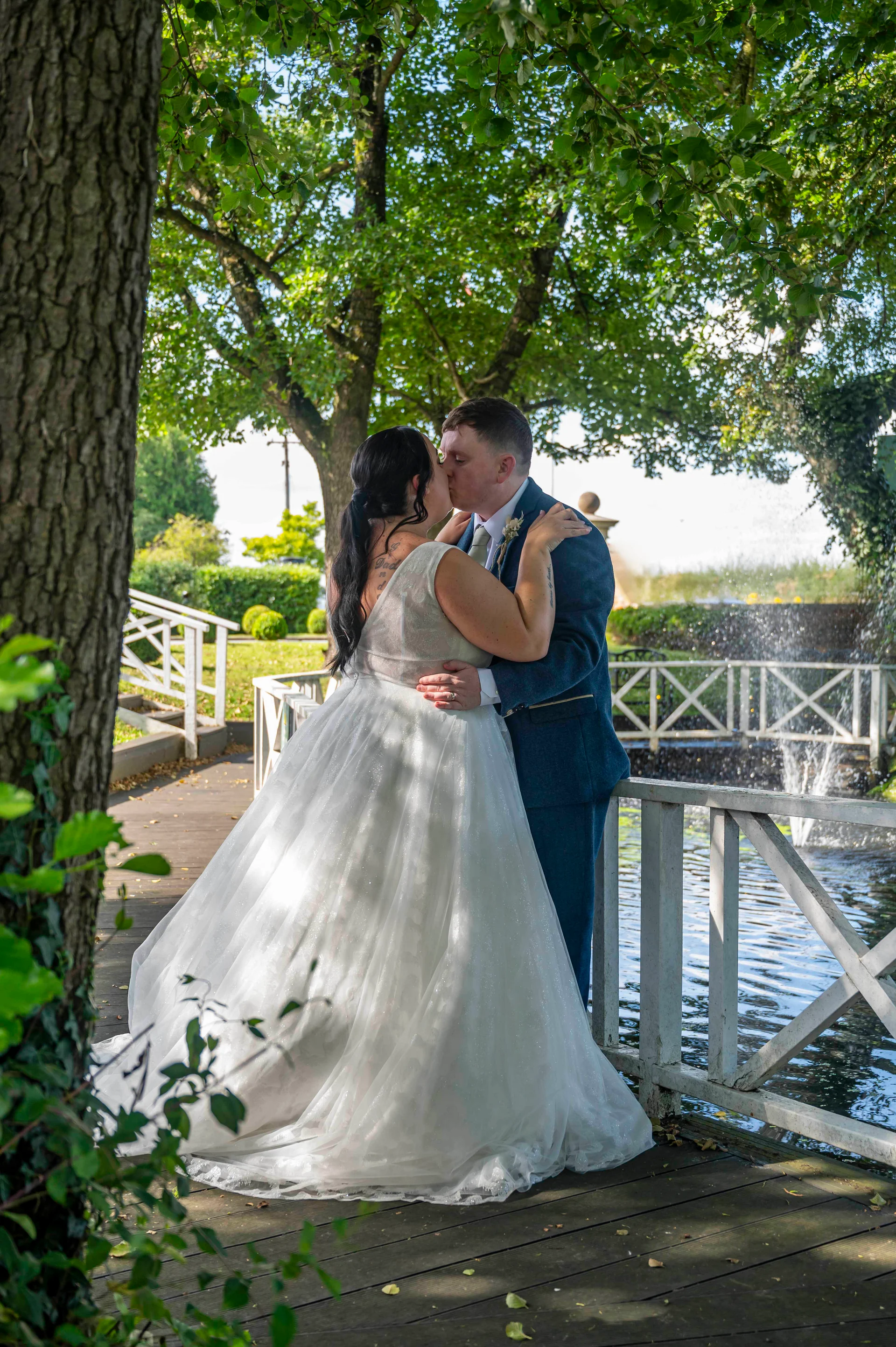 A newlywed couple shares a tender kiss on a wooden footbridge surrounded by lush trees and a scenic fountain.