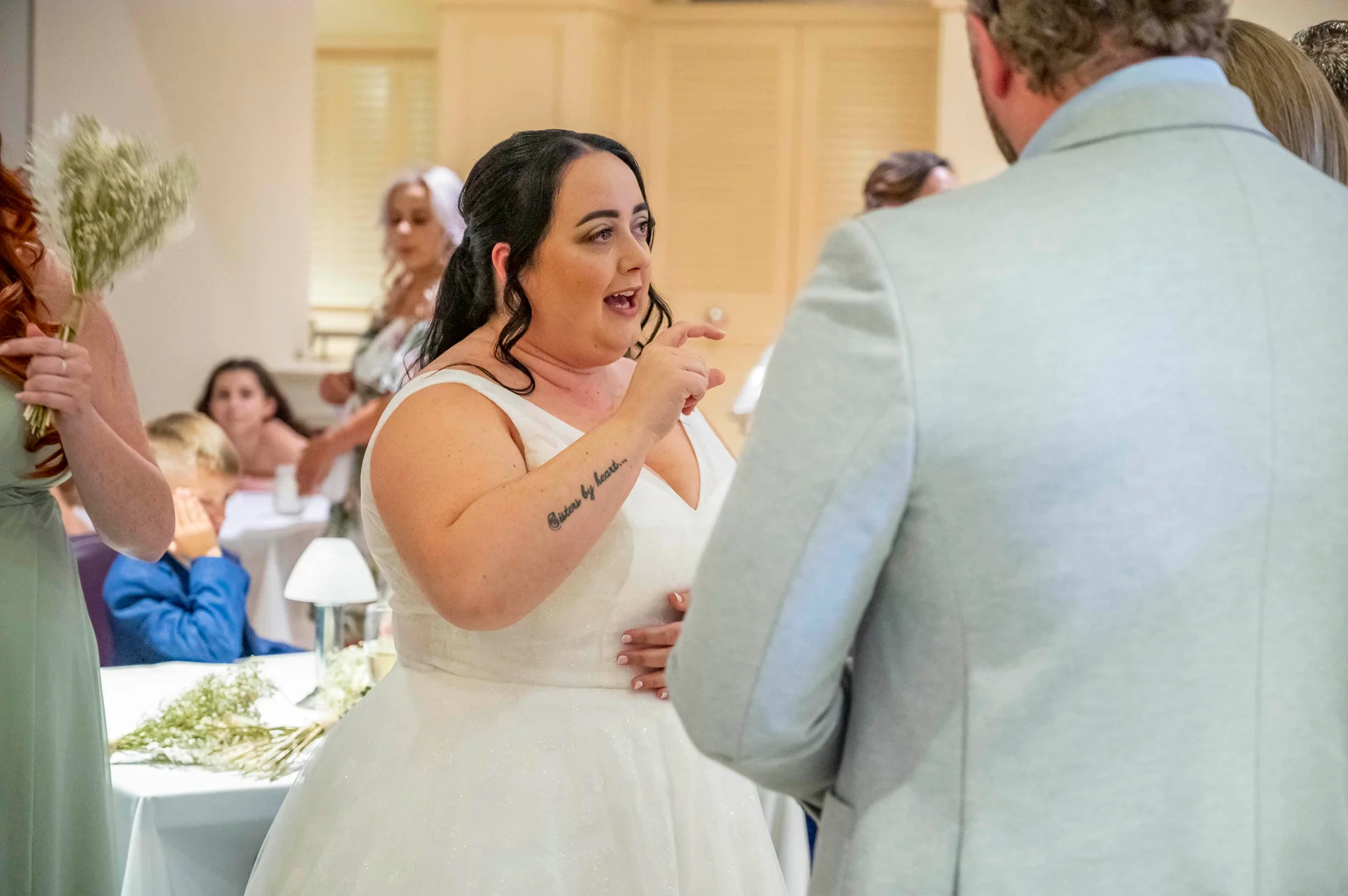 A joyful bride with an arm tattoo smiles and gestures while talking to a guest during the wedding reception.