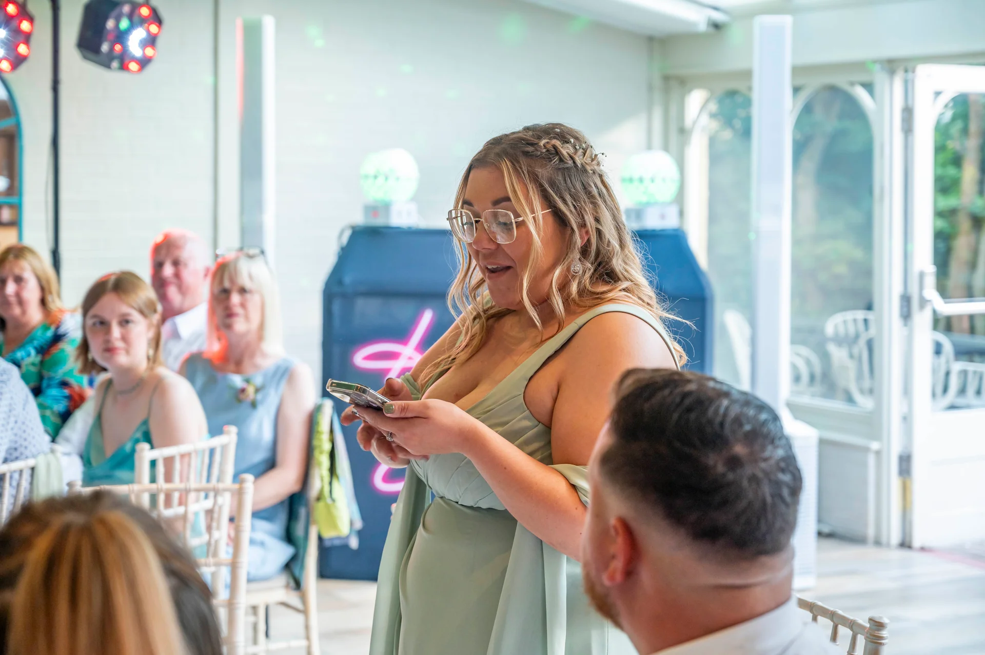 A bridesmaid in a sage green dress delivers a speech at her wedding reception, holding a smartphone while seated guests look on attentively.