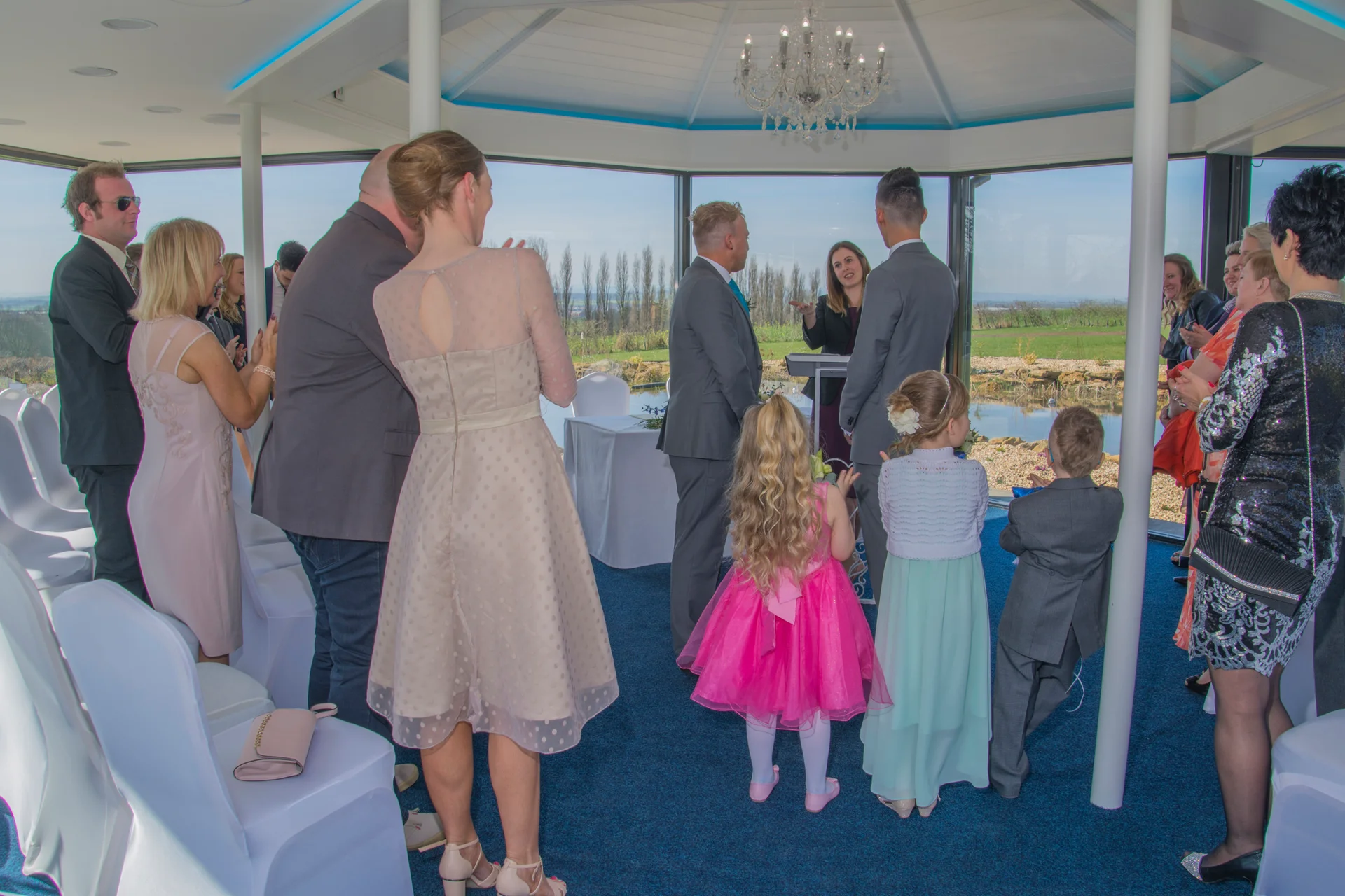 Family and wedding party members stand inside a sunlit, glass-walled pavilion during a civil wedding ceremony while looking towards the couple and celebrant.