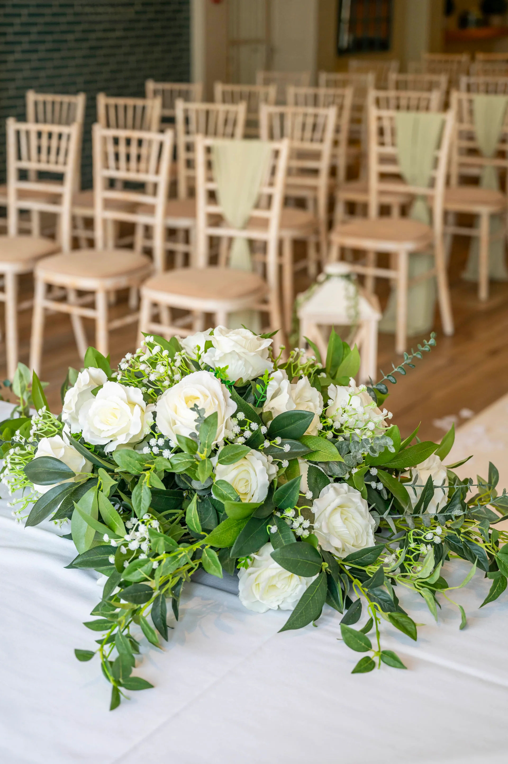 A lush floral arrangement of white roses, gypsophila, and eucalyptus sits atop a white linen-covered table in a bright wedding ceremony venue.
