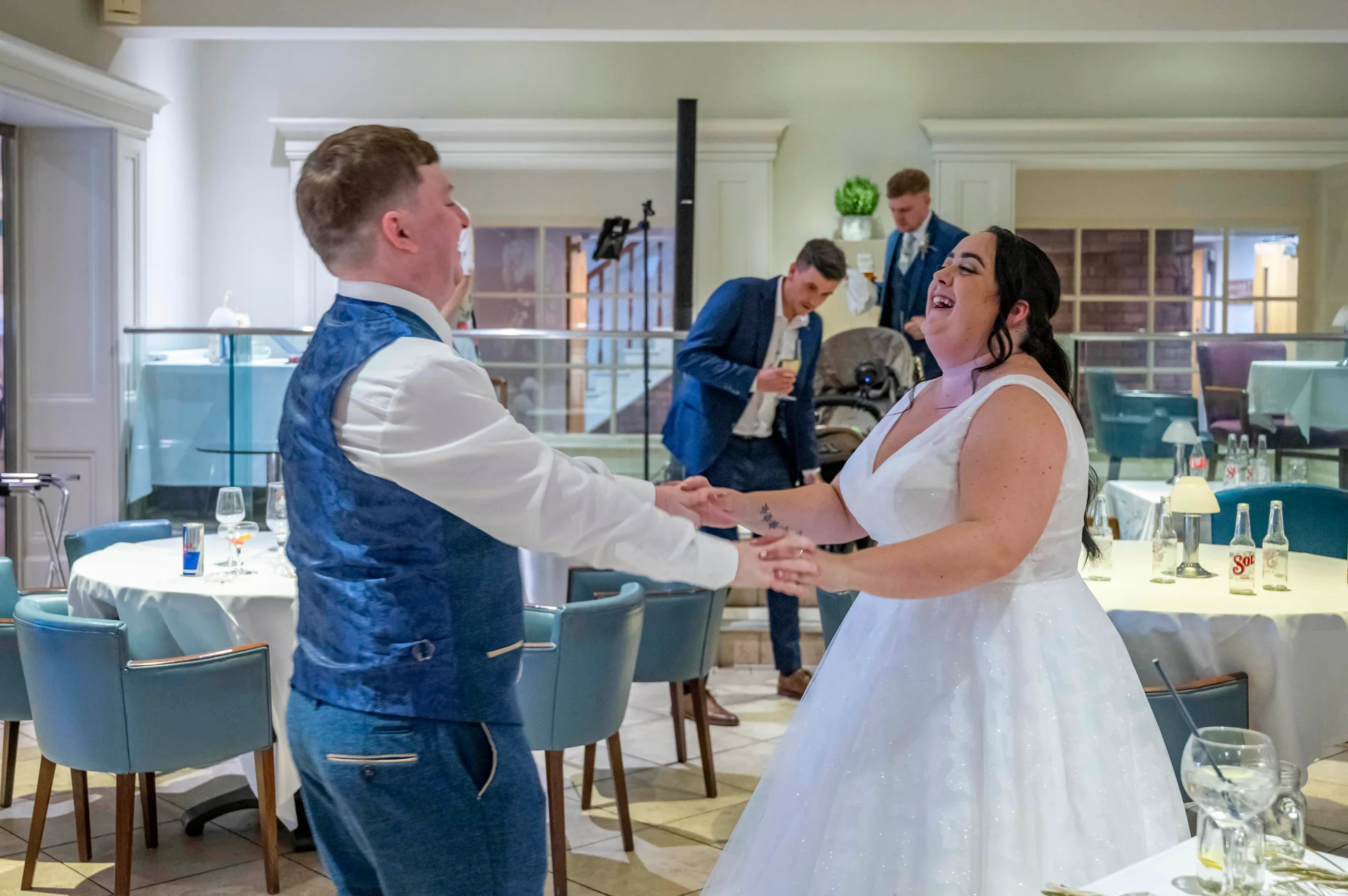 A newlywed couple share a joyful moment during their first dance, holding hands and laughing together in a bright reception room with blue chairs and dining tables.