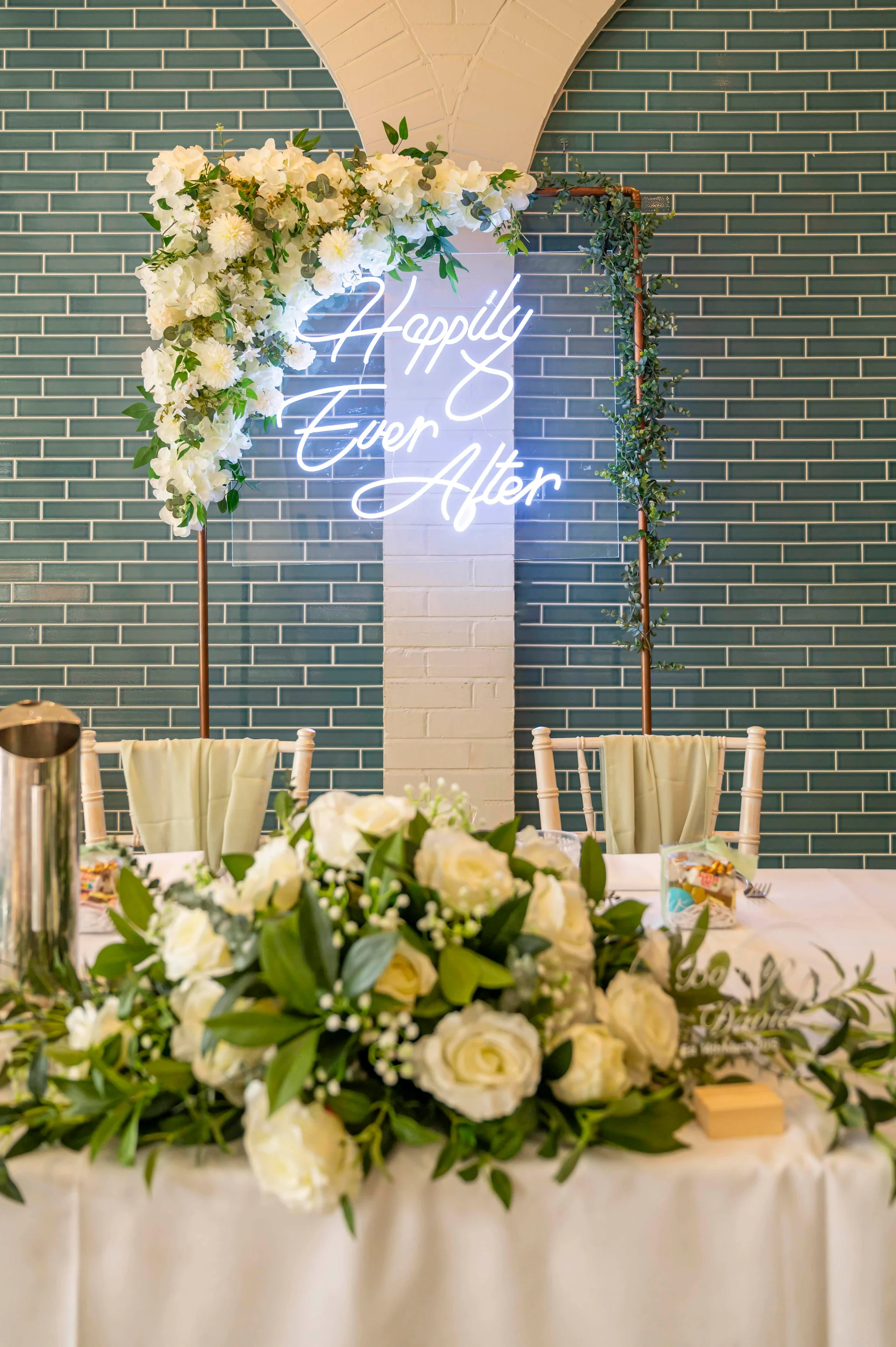A beautifully styled wedding top table features lush white rose and greenery arrangements, complemented by an illuminated neon sign reading Happily Ever After against a green tiled wall.
