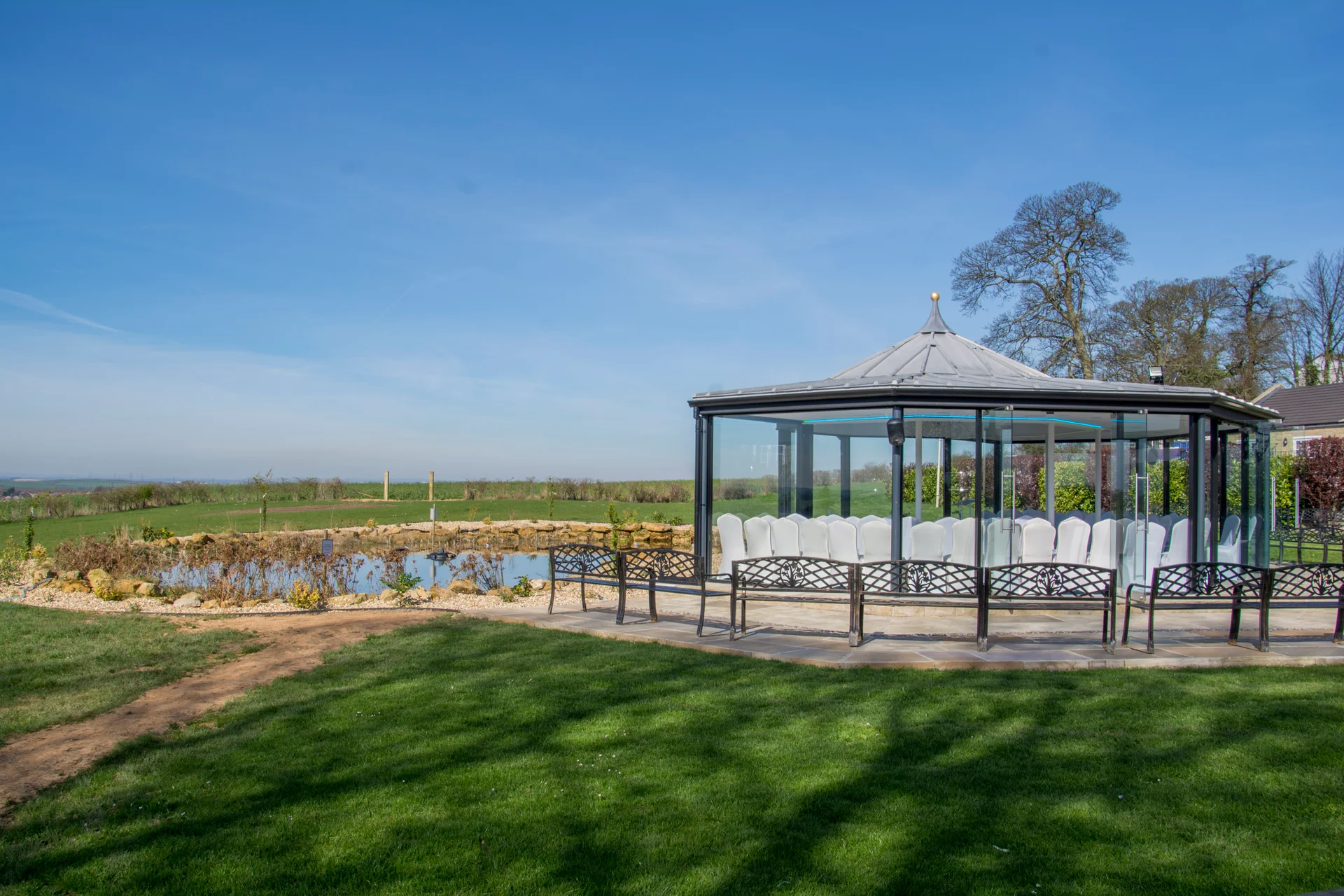 A modern glass gazebo wedding ceremony venue stands beside a tranquil pond under a clear blue sky in the countryside.