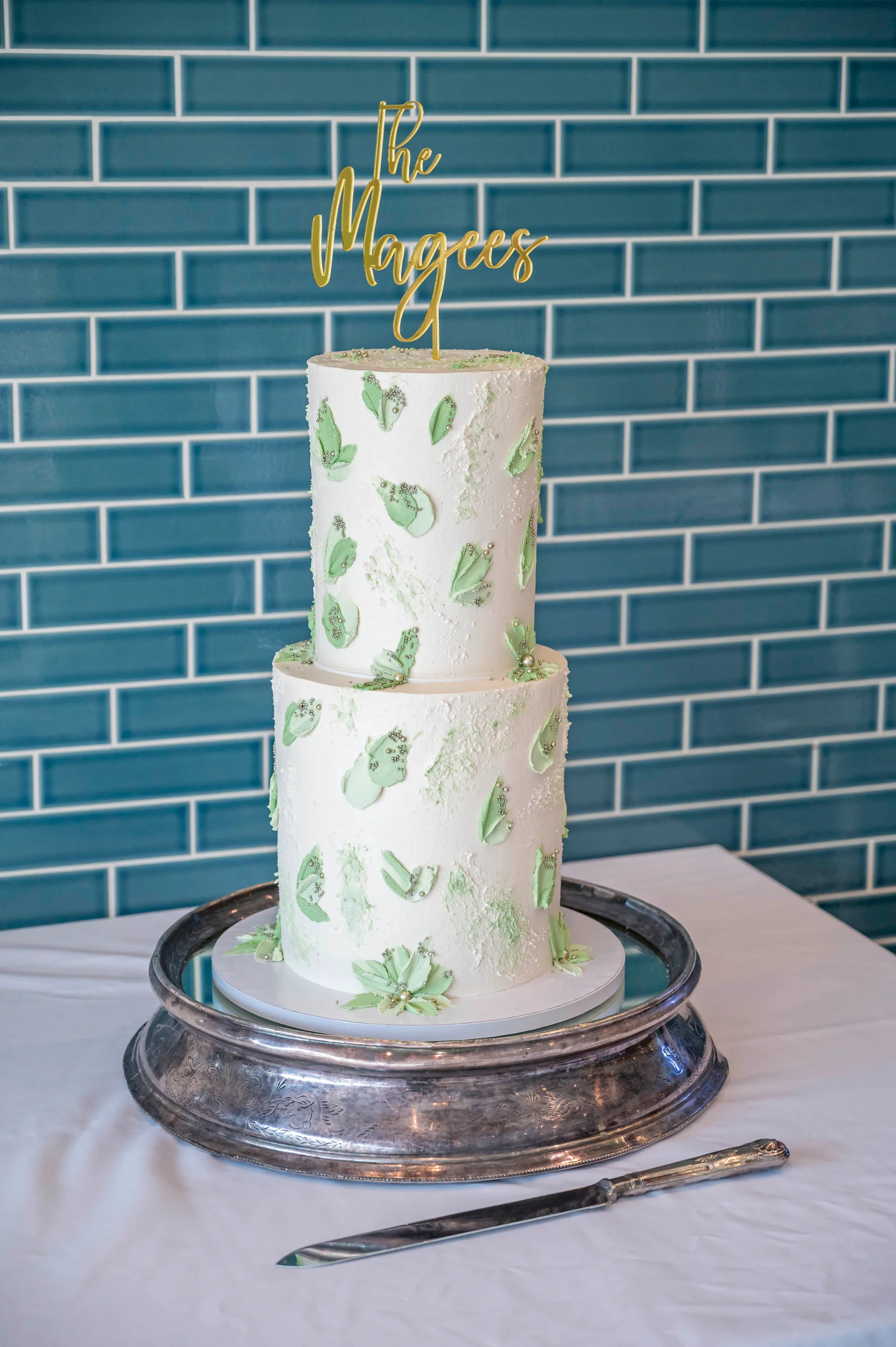 A elegant two-tier wedding cake featuring textured white buttercream and delicate sage green painted details rests on a silver tray against a teal tiled wall.