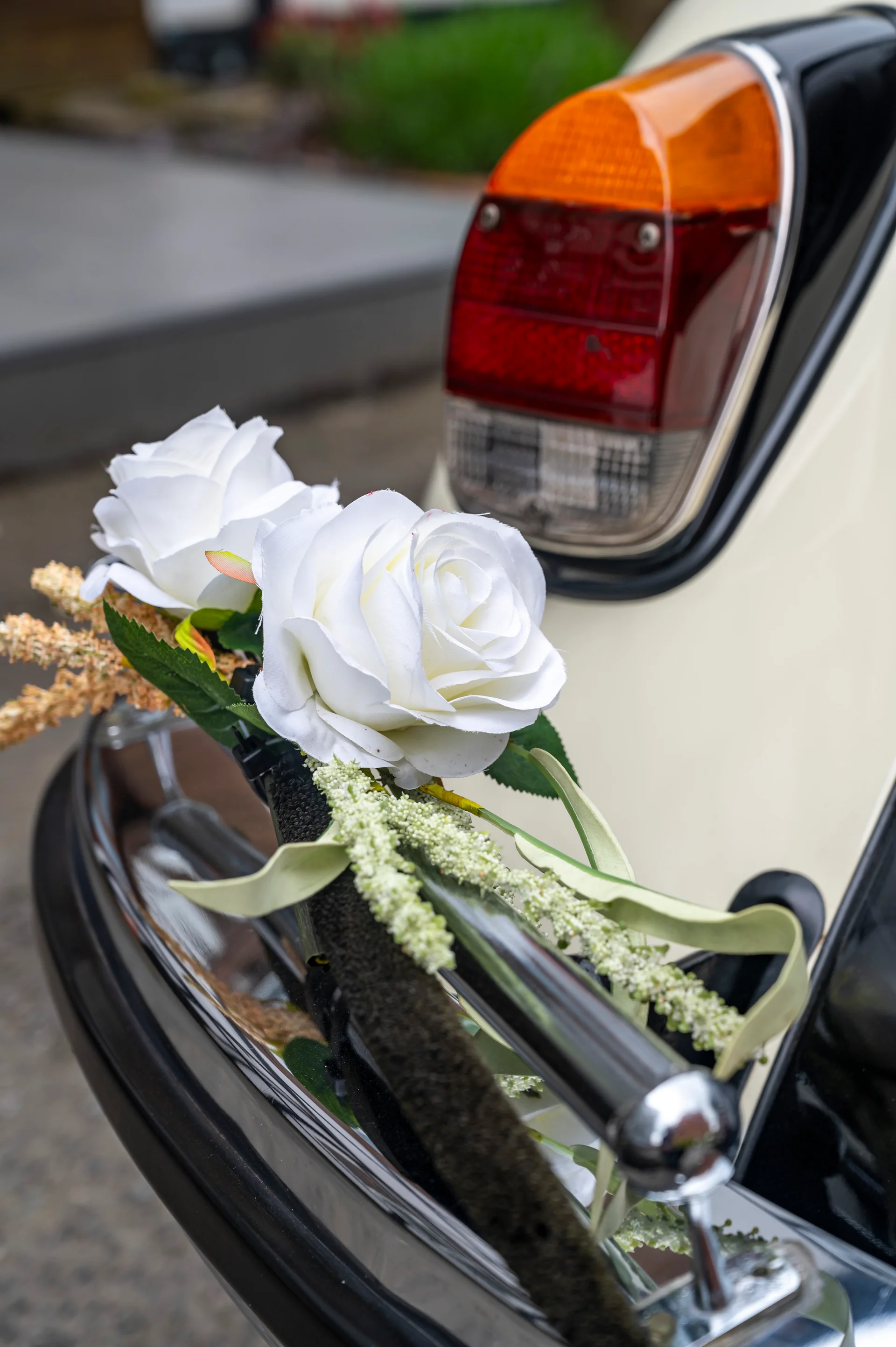 A close-up view of a vintage wedding car decorated with elegant white roses and greenery tied to the bumper near the rear light.
