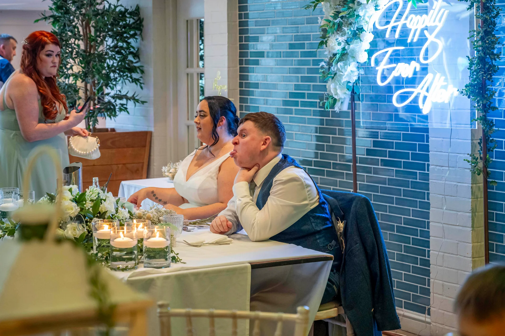A groom pulls a silly face at the top table beside his bride during the wedding breakfast, with a glowing neon sign mounted on a blue tiled wall behind them.