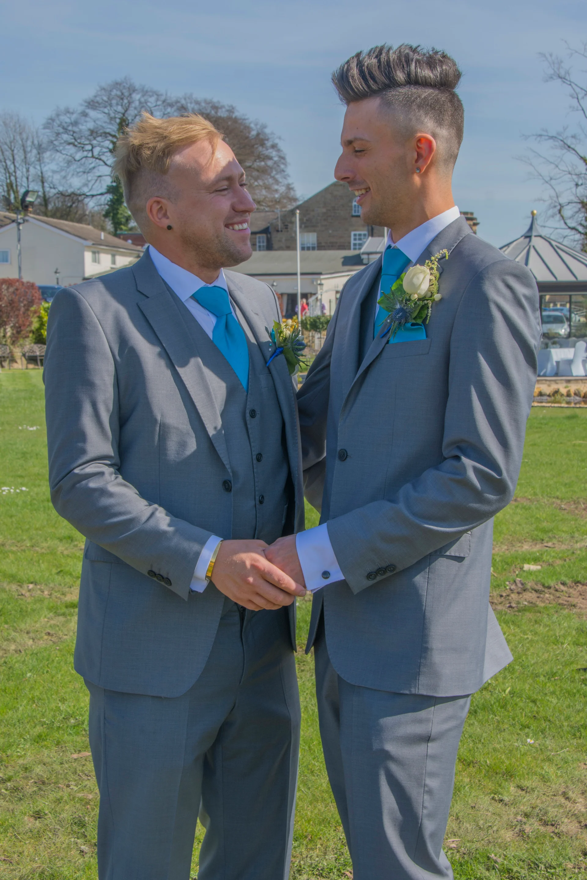 Two grooms in matching grey suits and bright blue ties smile lovingly at each other while holding hands outdoors on a sunny day.