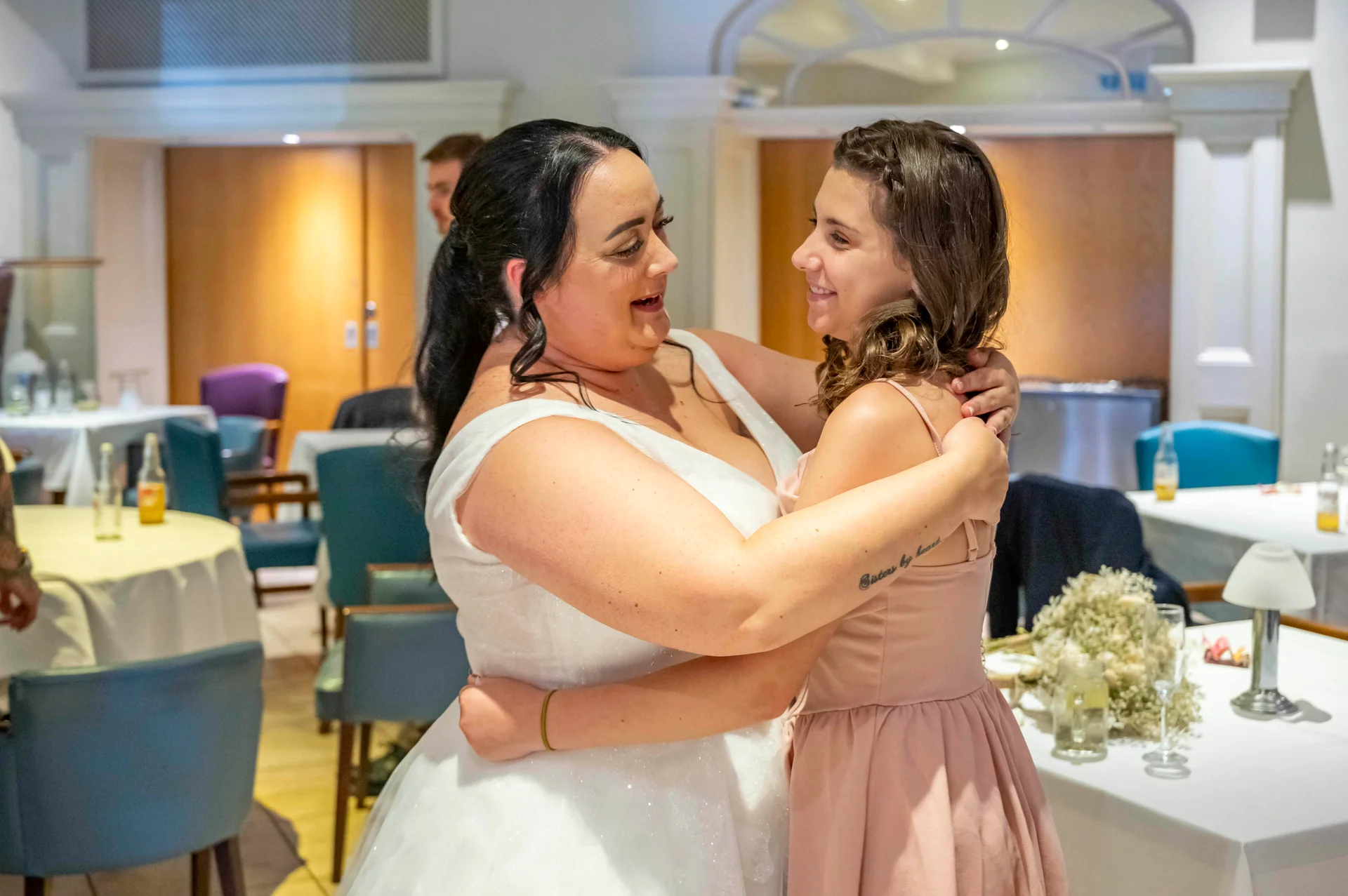Two smiling brides share a warm, joyful embrace during their wedding reception in a hotel dining room.