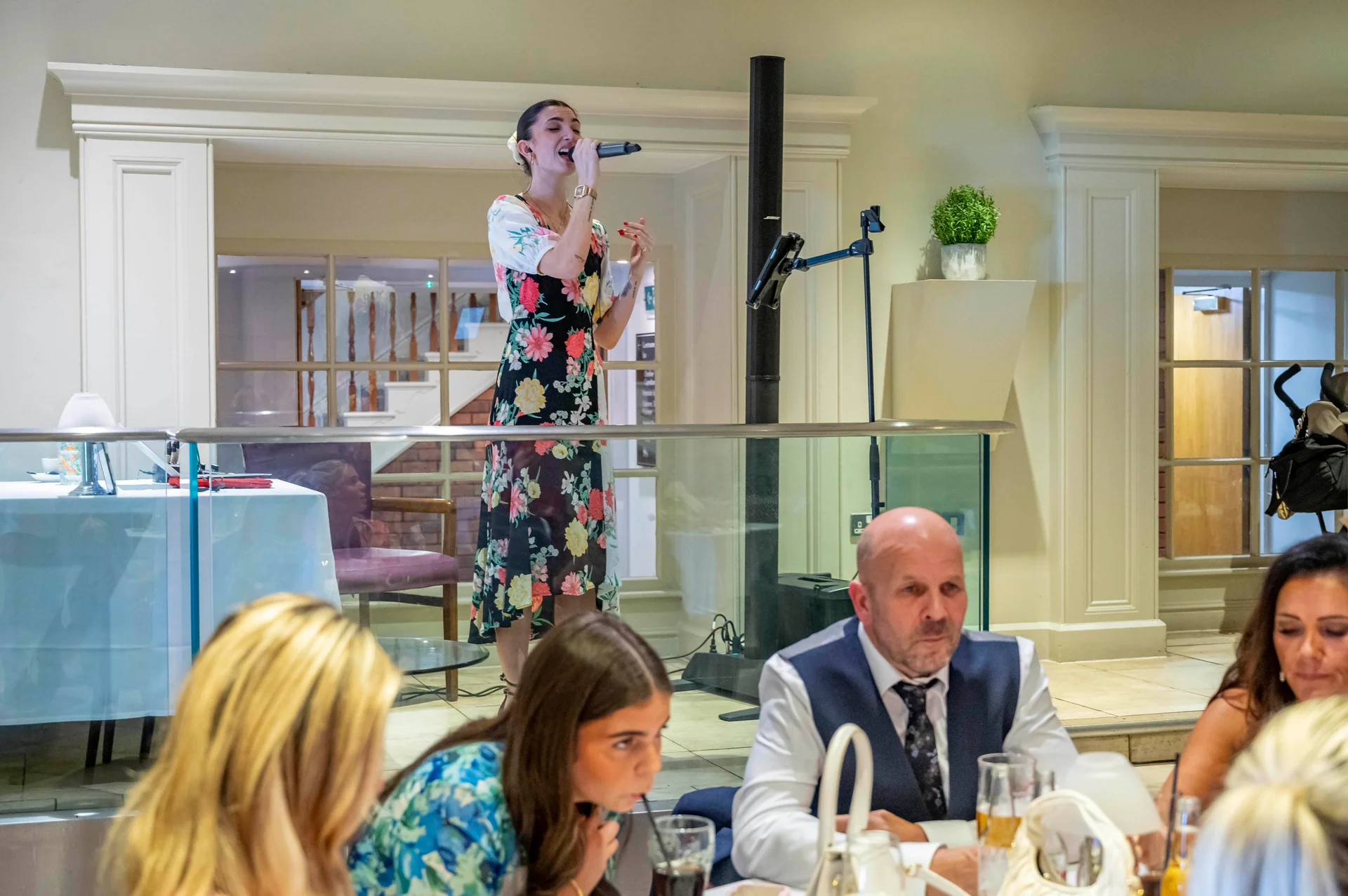A female vocalist performs with a microphone on a raised platform during a wedding reception while guests sit at tables below.