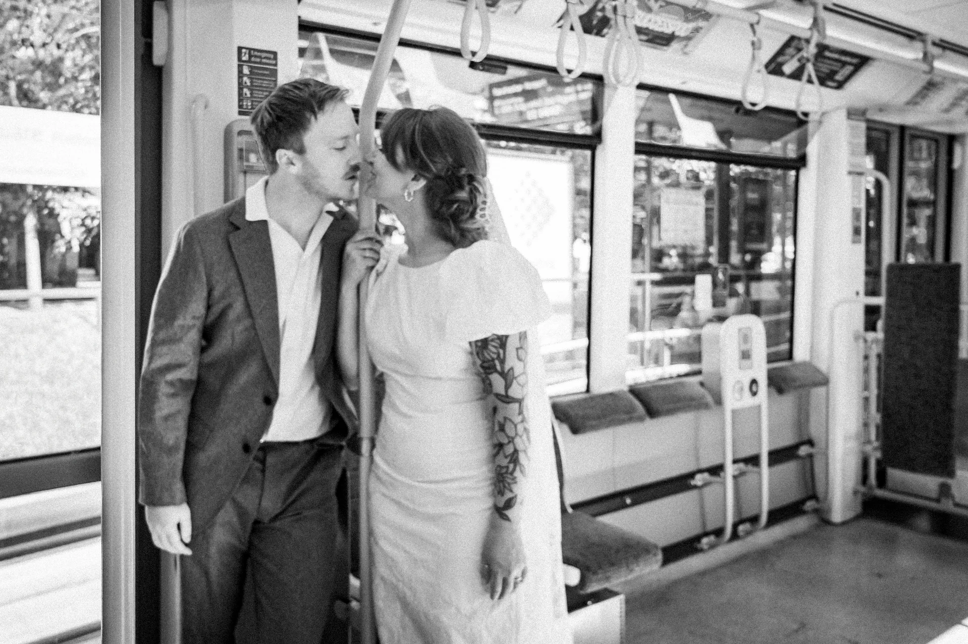 A newlywed couple shares an intimate kiss inside a public transport tram during their wedding day, captured in a candid black and white style.
