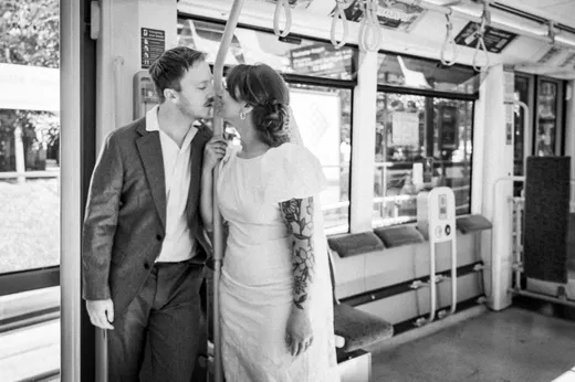 A newlywed couple shares an intimate kiss inside a public transport tram during their wedding day, captured in a candid black and white style.