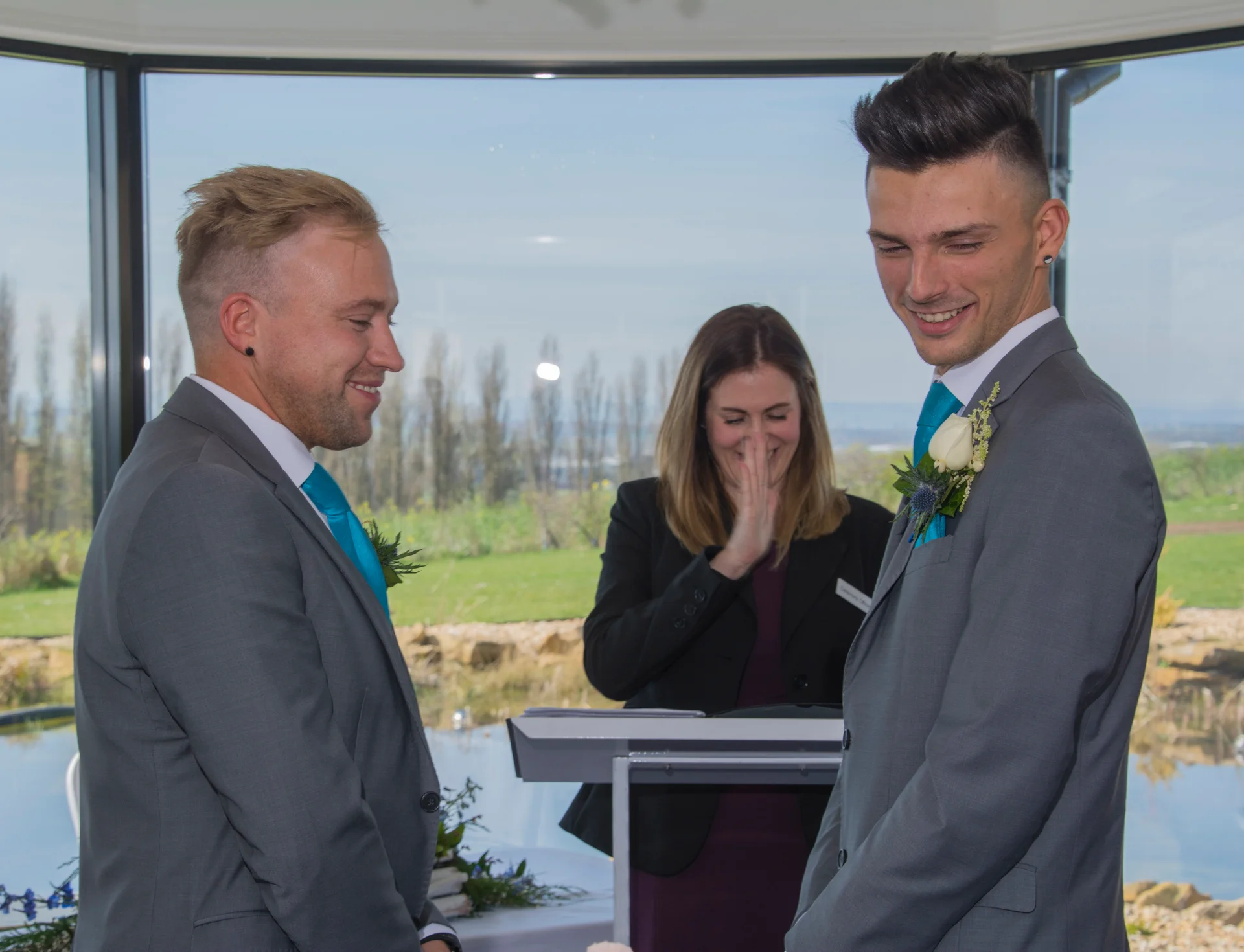 Two grooms in matching grey suits and turquoise ties smile joyfully during their civil wedding ceremony while an officiant looks on in the background.