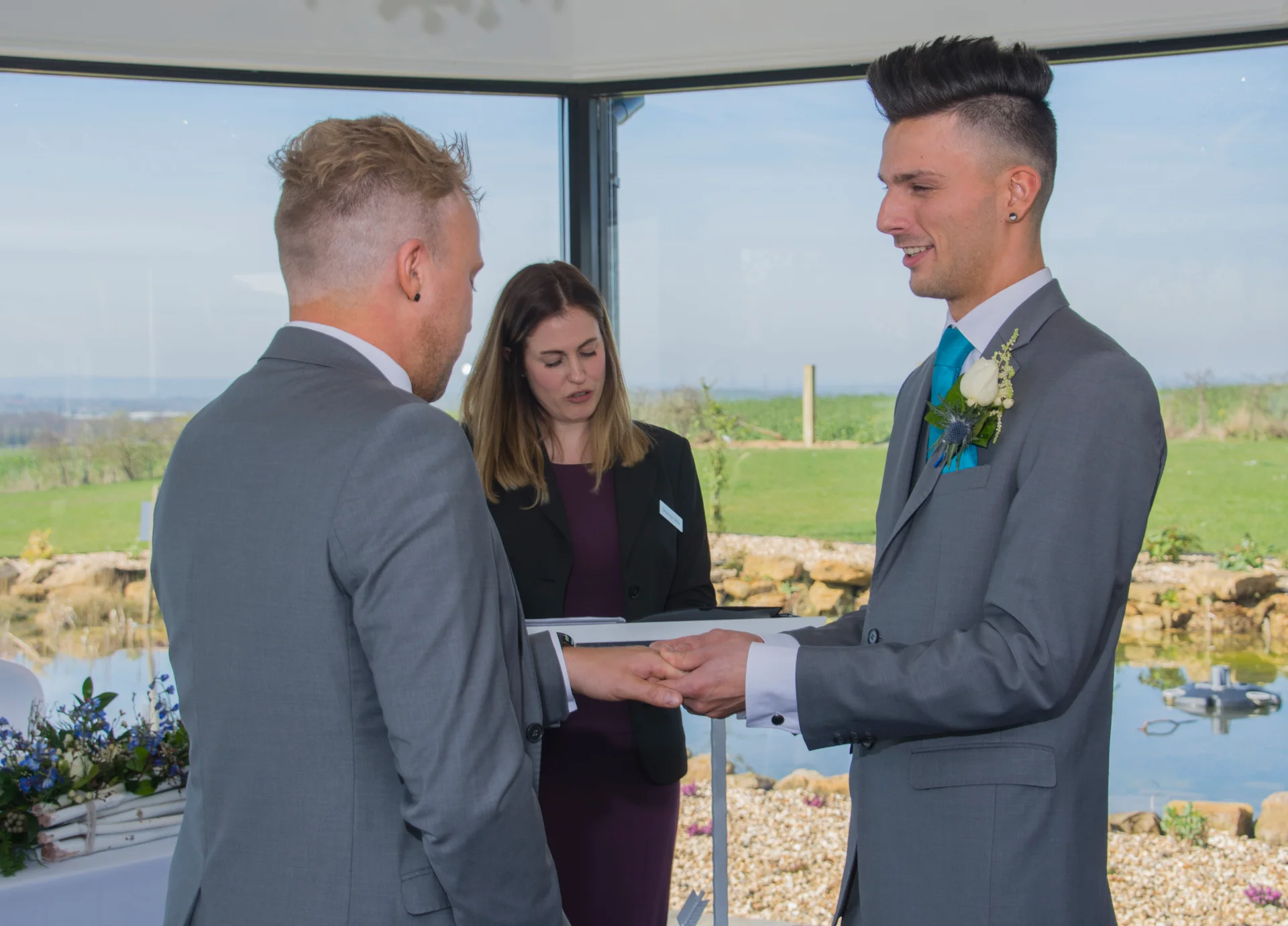 Two grooms exchange rings during an intimate civil ceremony officiated by a celebrant in a bright, light-filled room overlooking a scenic outdoor landscape.