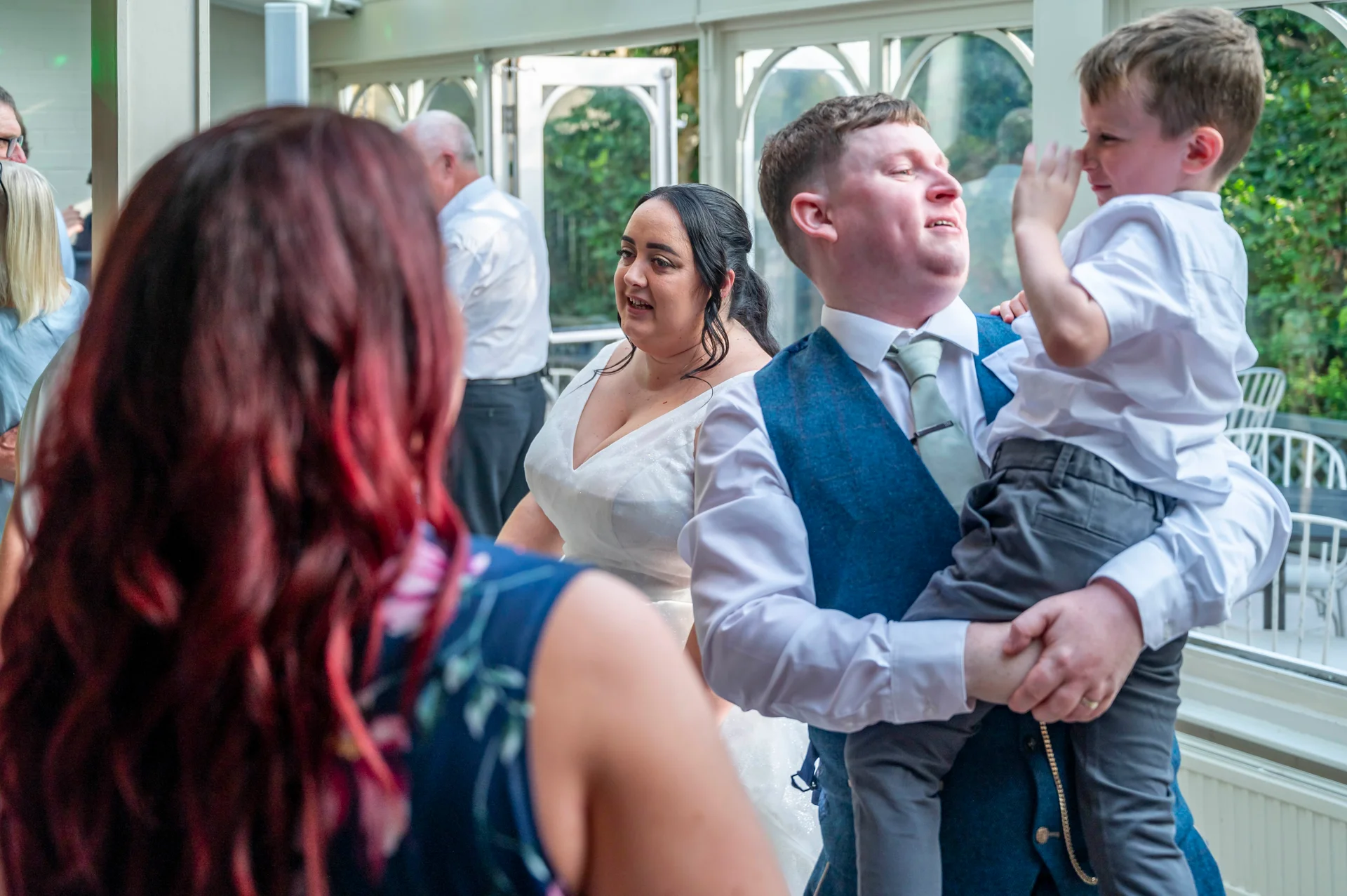 A groom holding a young boy in his arms while standing with the bride inside a light-filled conservatory venue.