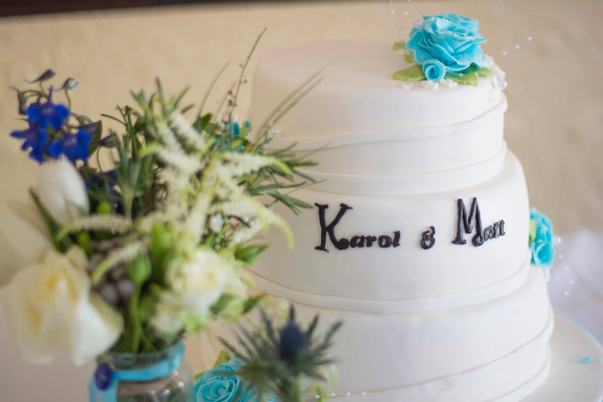 A three-tier white fondant wedding cake adorned with blue sugar flowers and personalised lettering stands next to a floral arrangement featuring blue delphiniums and thistles.