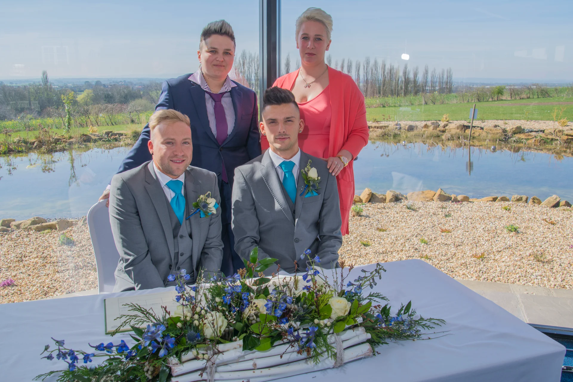 A bridal party poses together behind a signing table decorated with blue and white flowers, with a scenic lake view visible through large windows.