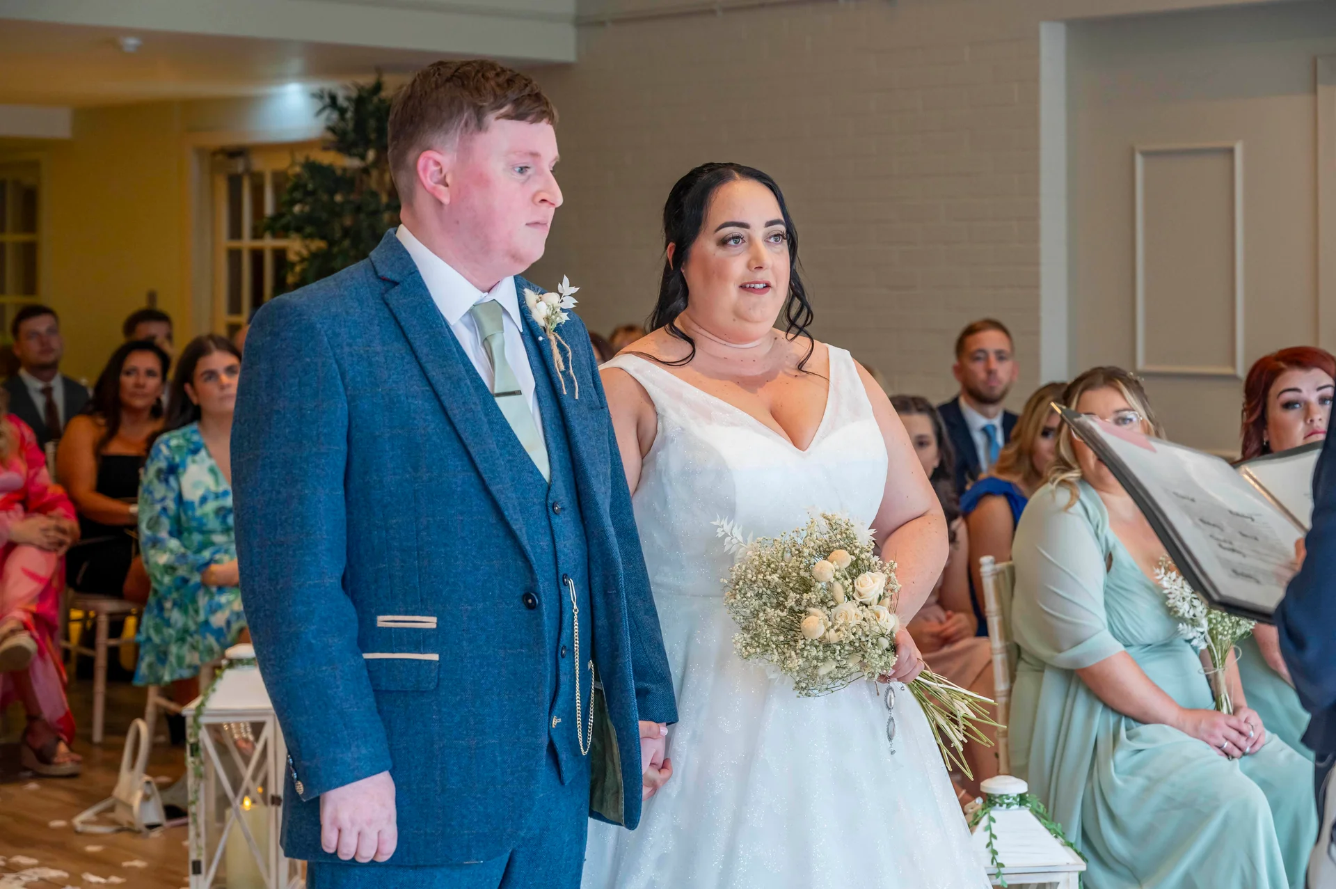 A bride in a white sparkling a-line gown holding a bouquet and a groom in a blue tweed suit stand together during their indoor wedding ceremony.