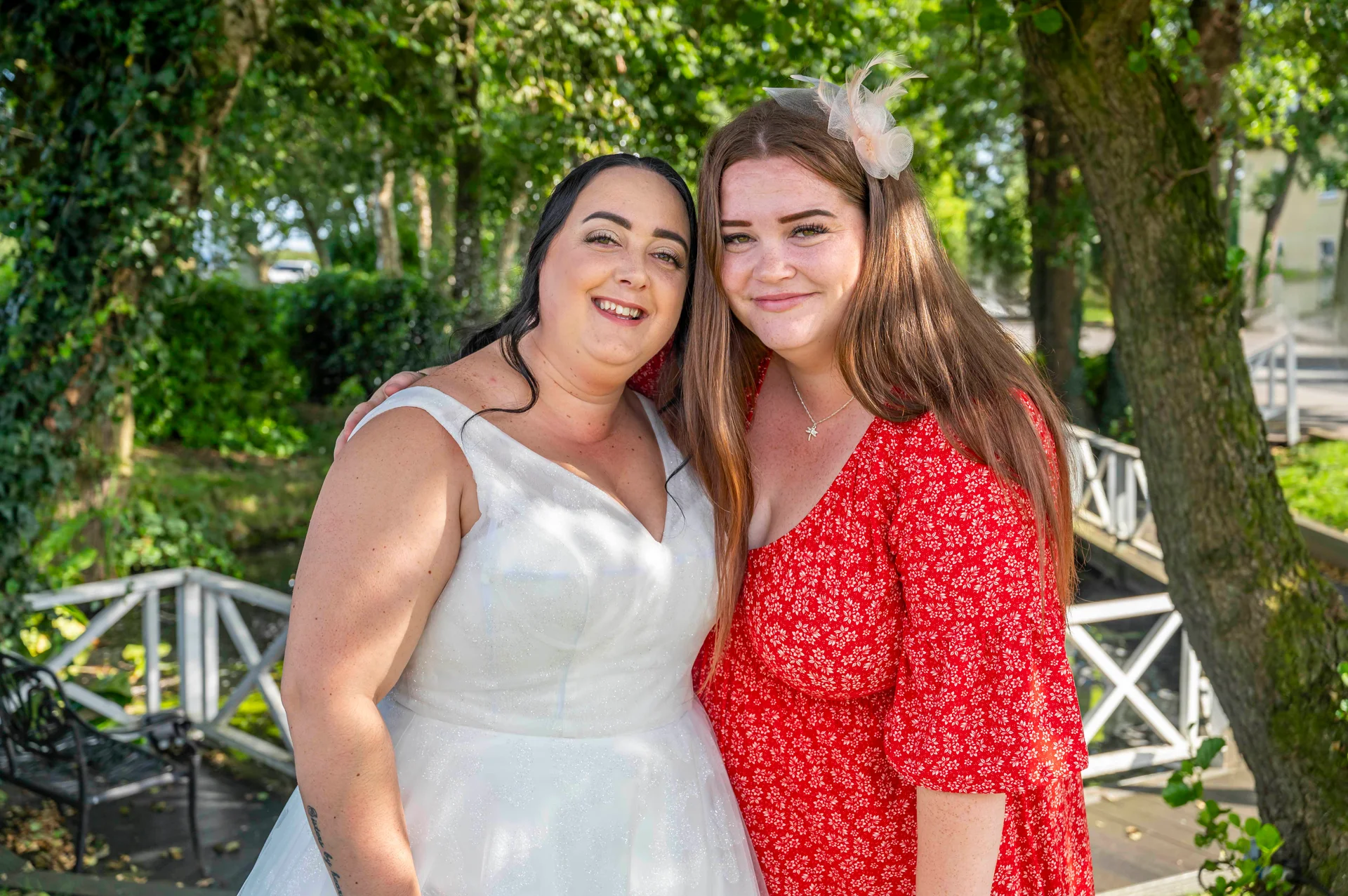 Two smiling brides embrace in an outdoor garden setting, with one wearing a white sparkle wedding dress and the other in a patterned red dress with a delicate fascinator.