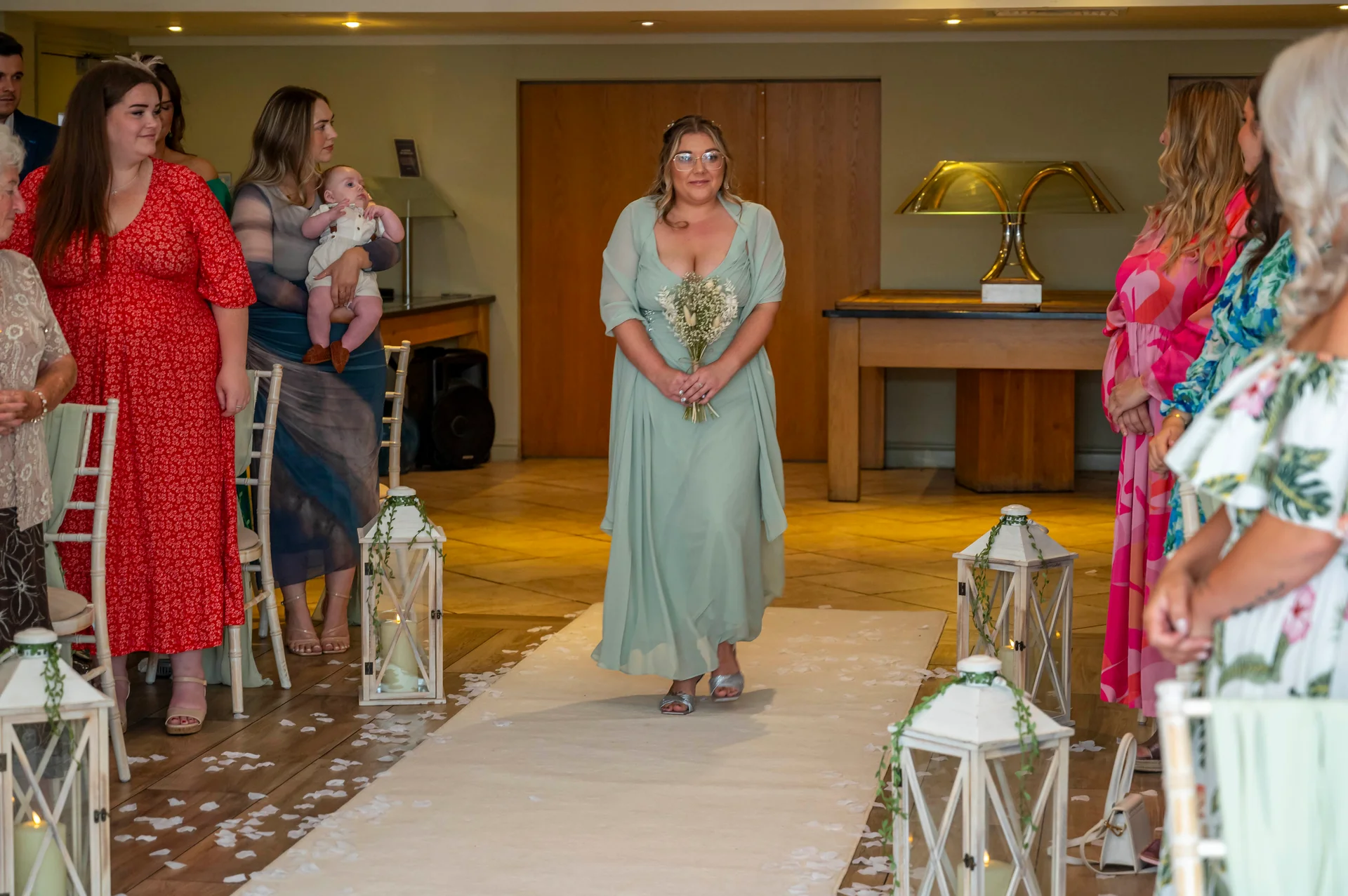 A smiling bride wearing a sage green dress and holding a bouquet walks down the white-carpeted aisle lined with lanterns.