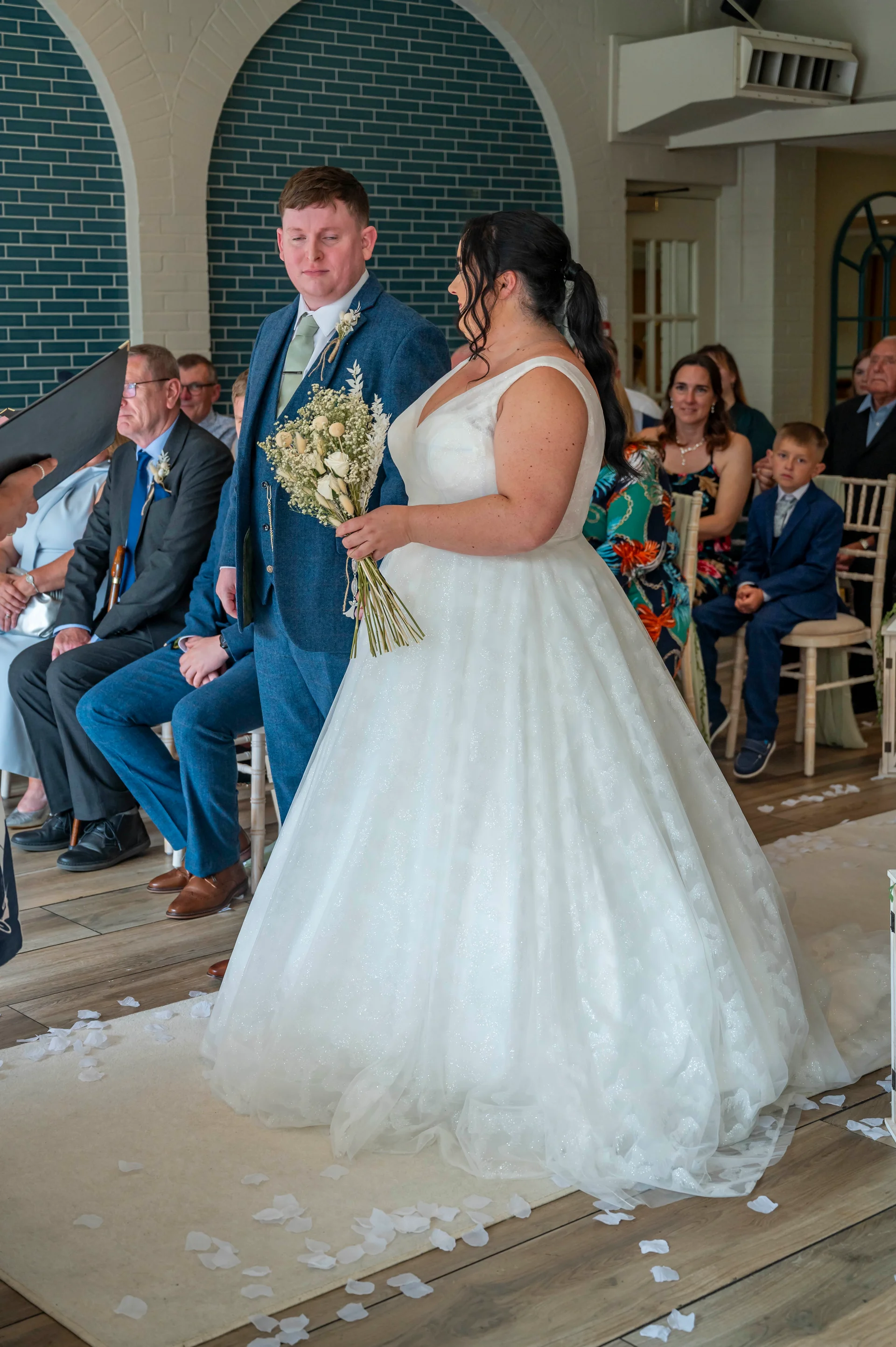 A bride in an a-line glitter tulle gown and a groom in a blue suit stand together at the front of a light-filled ceremony room with seated guests.