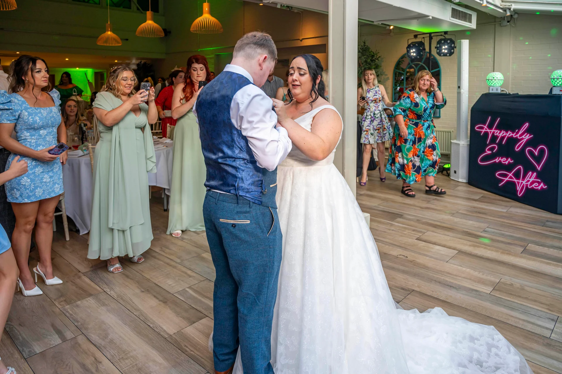 A newlywed couple shares an emotional first dance together on a wooden floor in a reception venue, watched by smiling guests and a glowing neon sign.