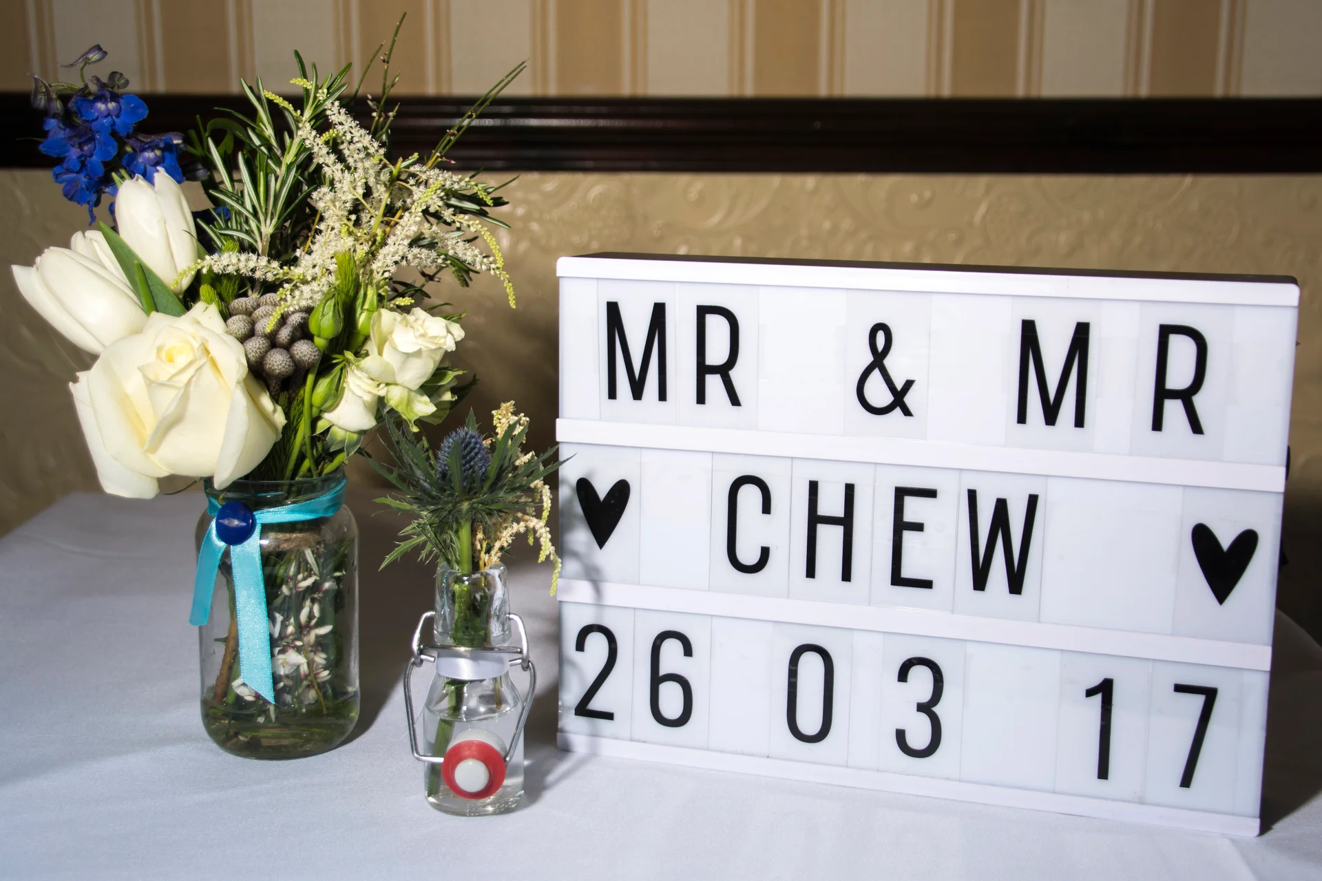 A light box reading Mr & Mr Chew with the wedding date rests beside a rustic floral arrangement in a glass jar on a white tablecloth.