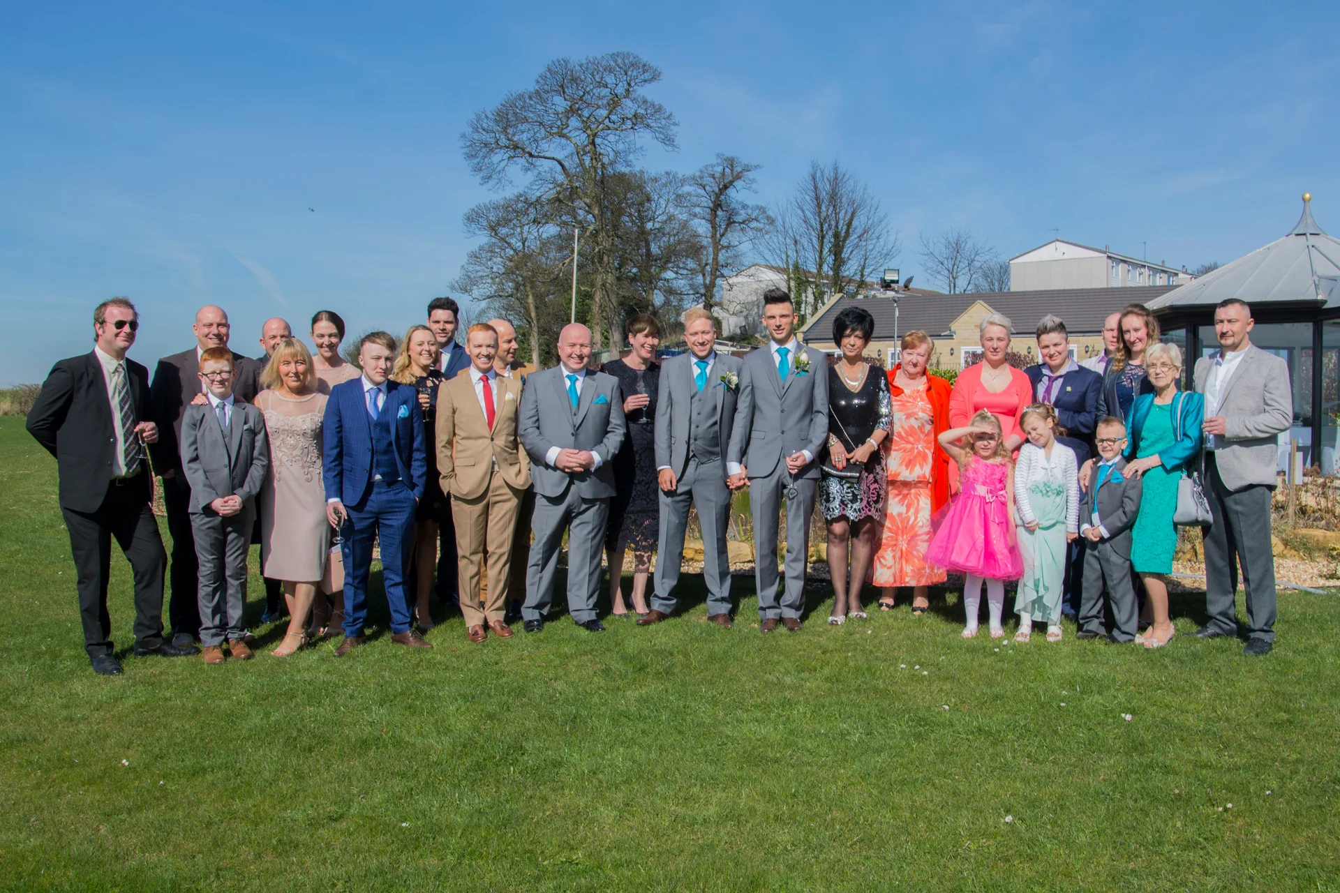 A large wedding party and family members pose outdoors on a sunny green lawn under a clear blue sky.