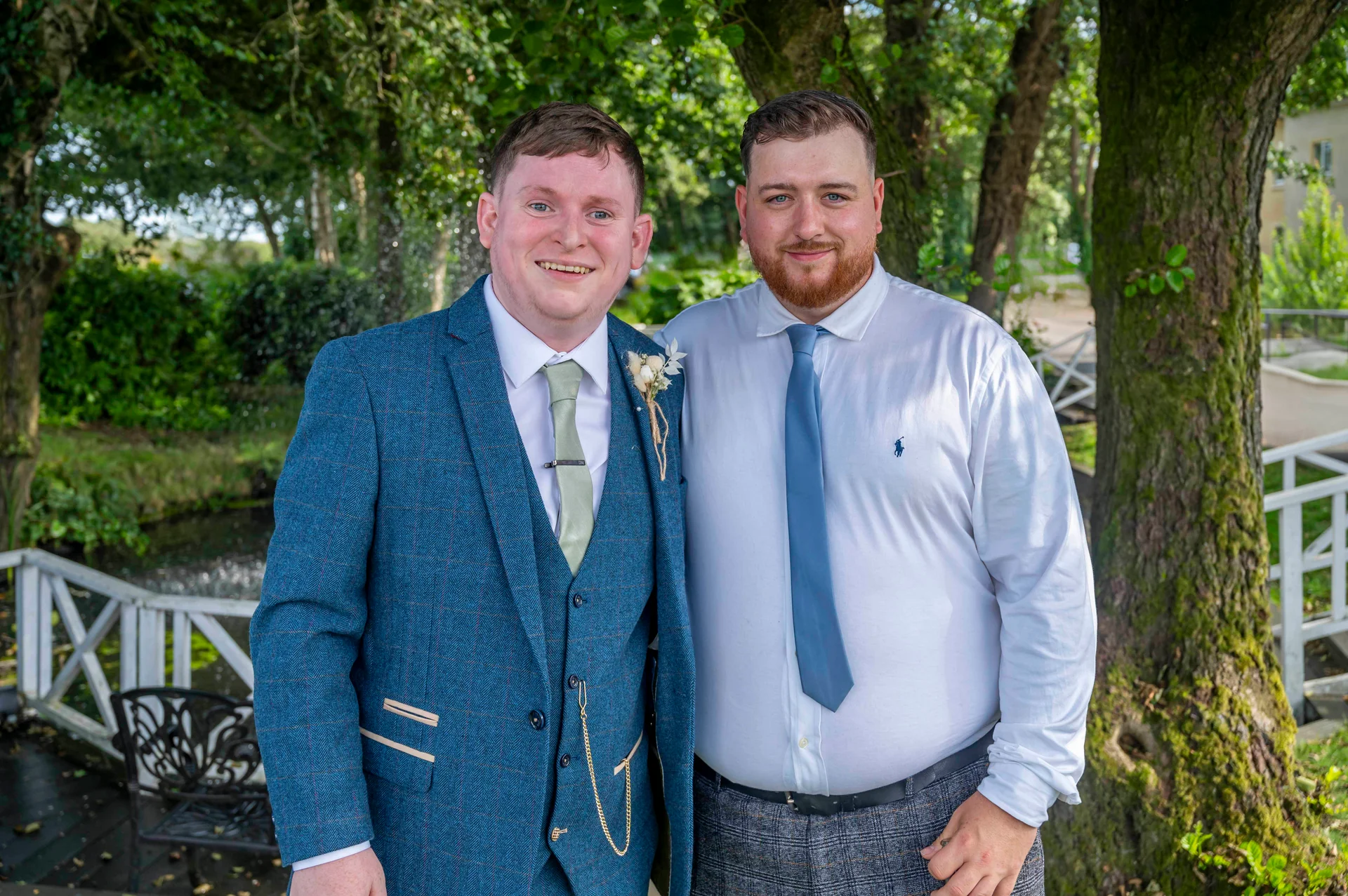 Two grooms stand side by side outdoors in a lush garden setting, one wearing a blue tweed three-piece suit with a pocket watch and the other in a crisp white shirt and tie.
