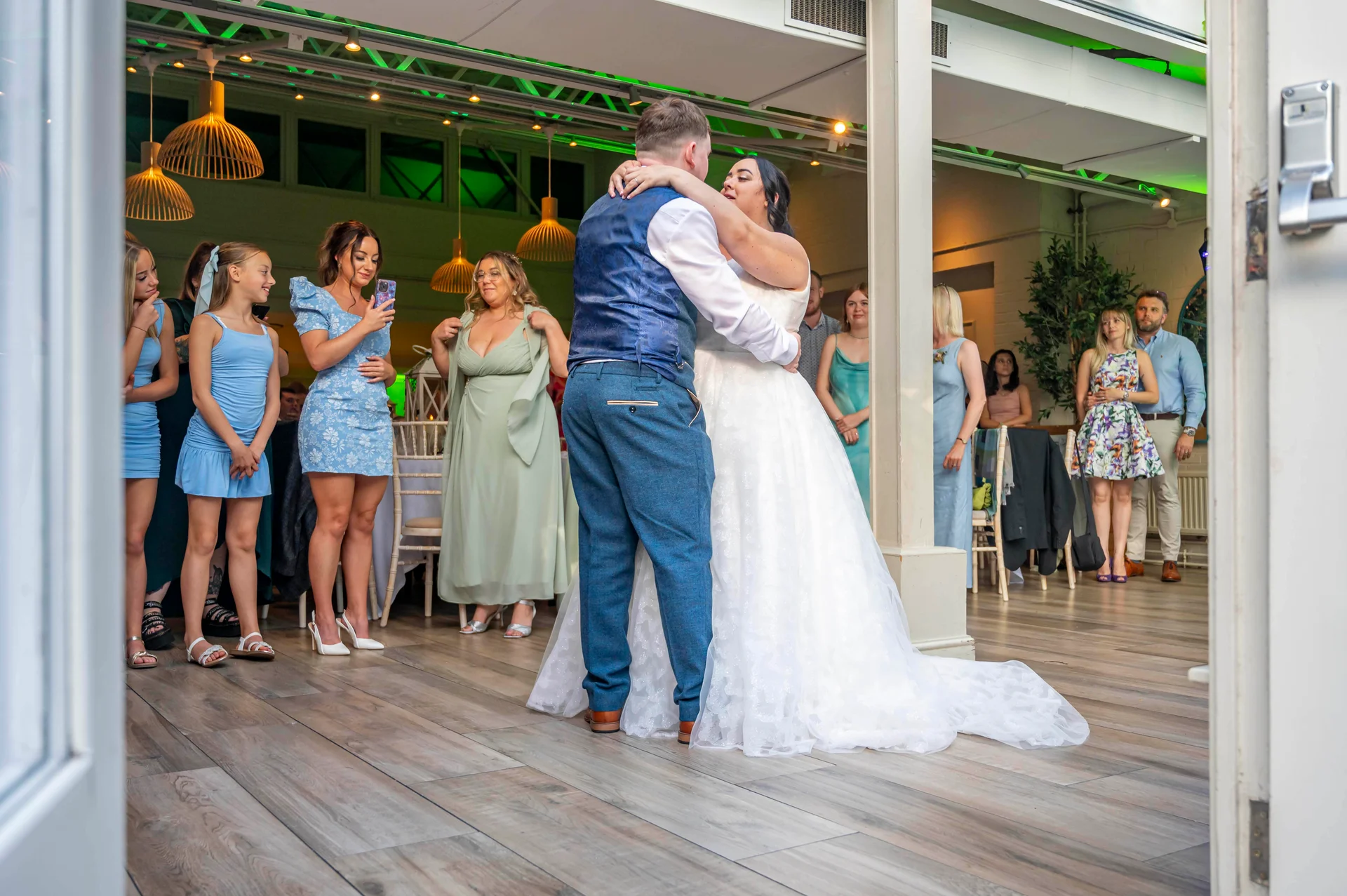 Newlyweds share their first dance on a wooden floor in a modern venue while guests watch admiringly from the side.