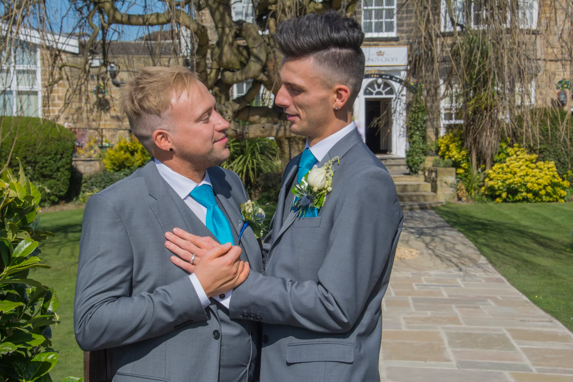 Two grooms in matching grey suits and bright blue ties lovingly hold hands and look into each other's eyes in the manicured gardens of a country house venue.