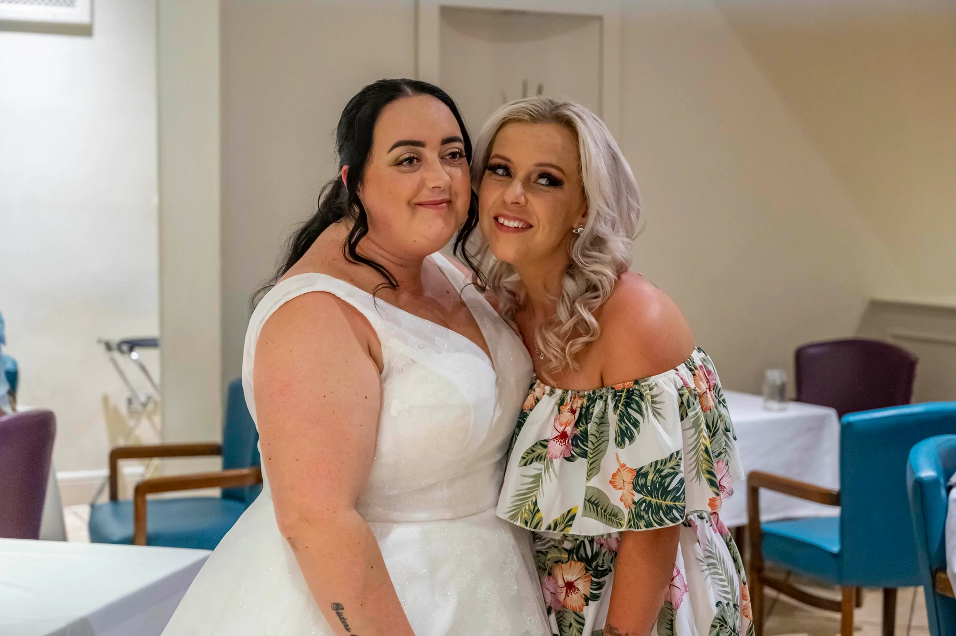 Two smiling brides embrace tenderly indoors, one wearing a sparkling white a-line gown and the other in a tropical floral print off-the-shoulder dress.