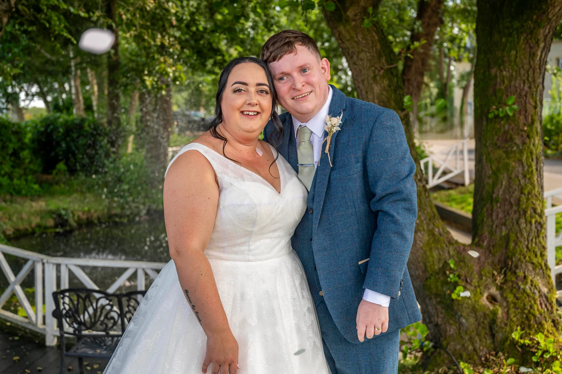 A smiling newlywed couple embraces outdoors near a tranquil pond, with falling confetti catching the sunlight during their wedding celebration.