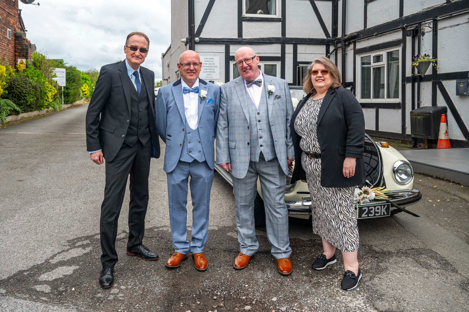 A group of four wedding guests and couple pose outdoors in front of a classic vintage car parked outside a building.
