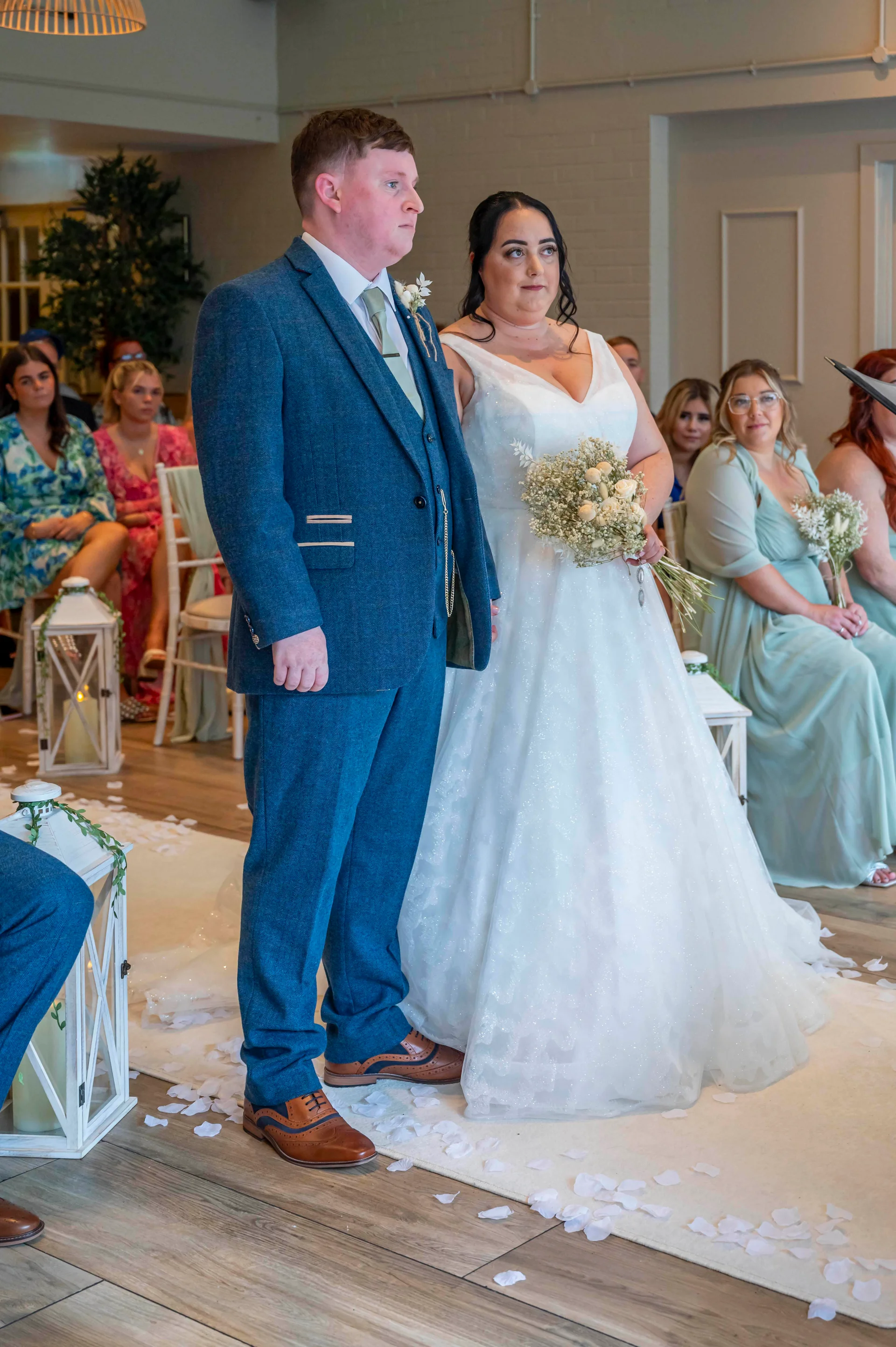 A bride in a sparkling a-line gown holding a posy bouquet stands beside her groom in a blue tweed suit during a civil ceremony.
