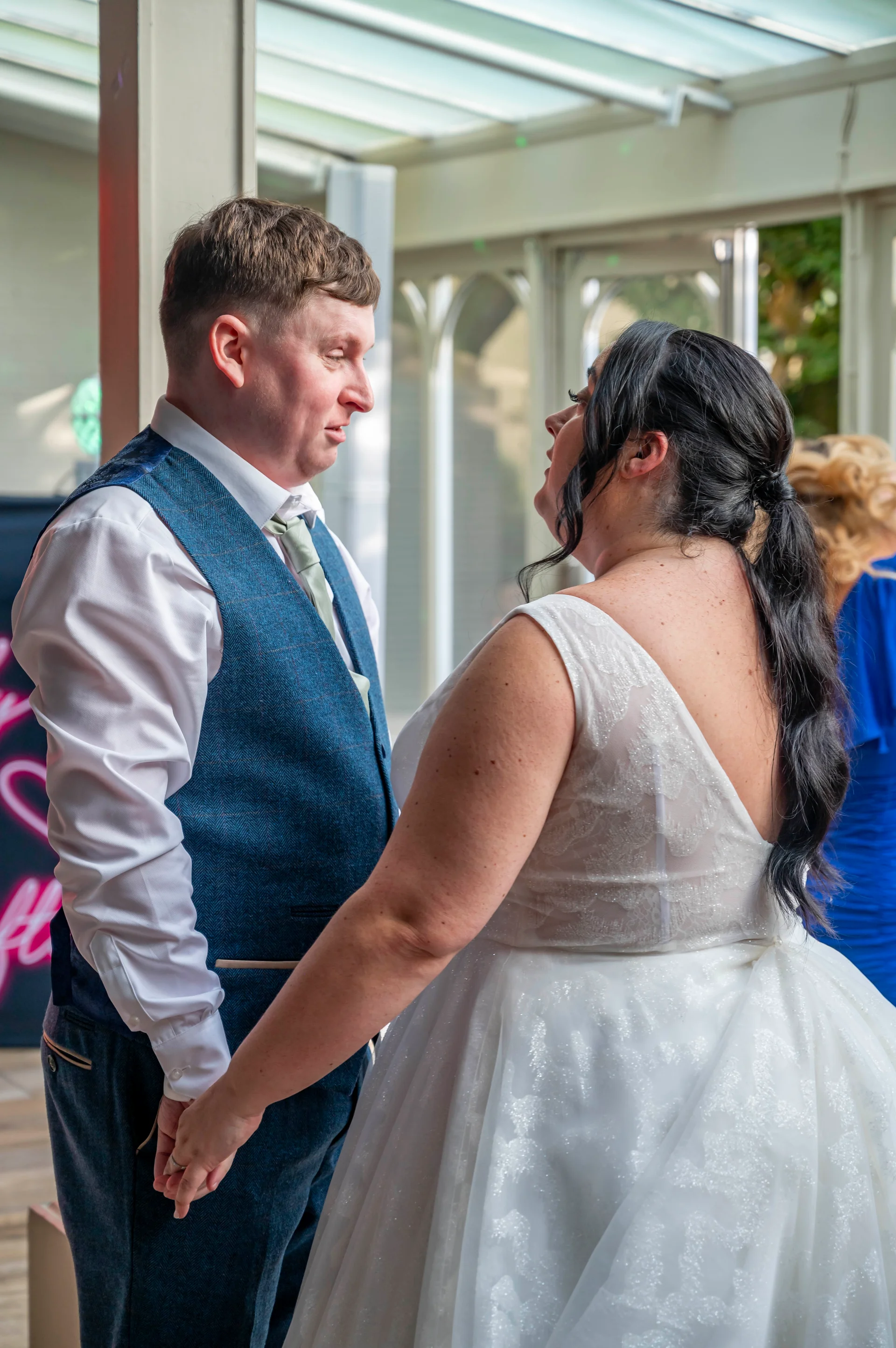 A bride and groom lovingly hold hands and look into each other's eyes inside a bright conservatory during their wedding reception.