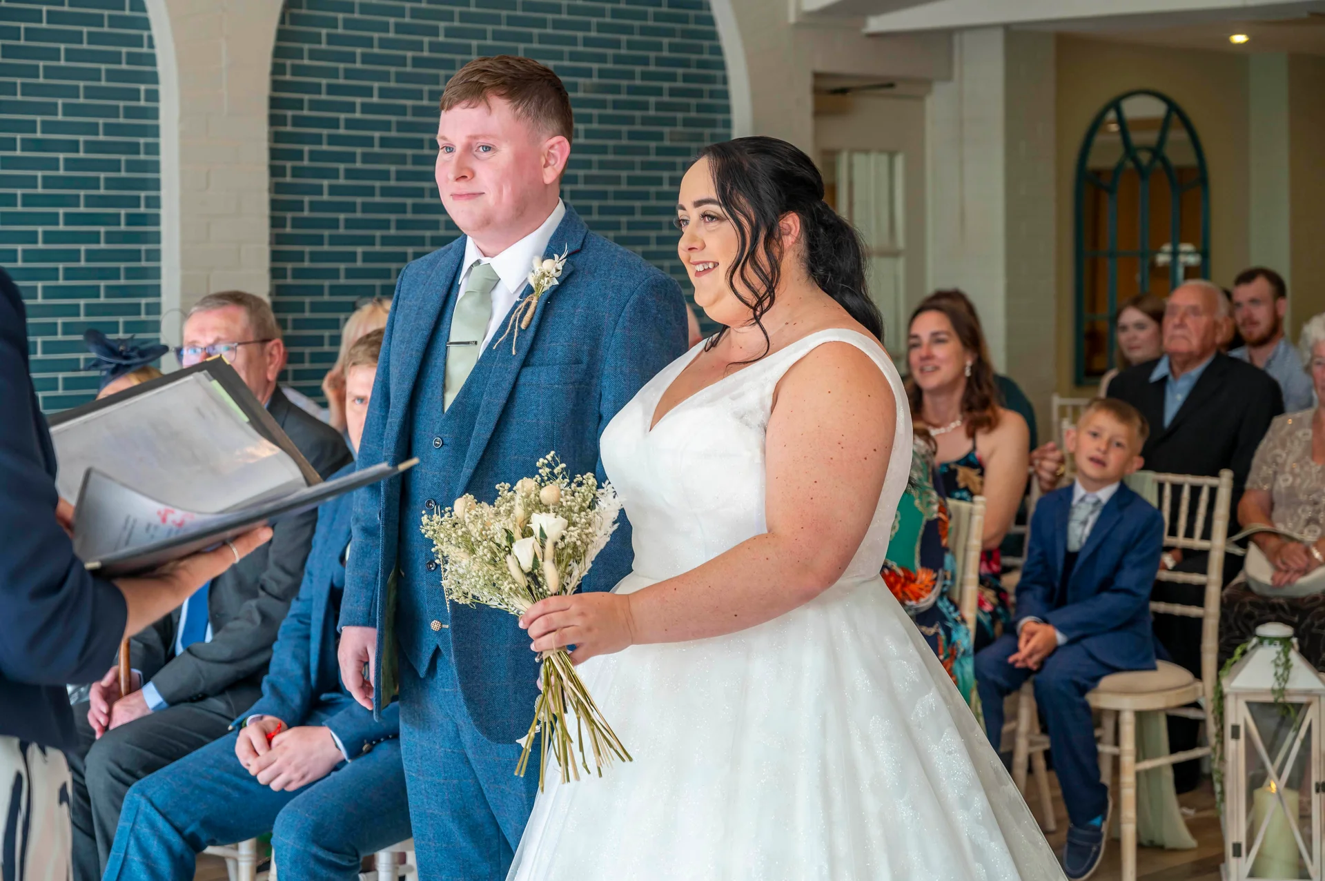 A smiling bride holding a dried floral bouquet and a groom in a blue tweed suit stand together during their indoor civil ceremony.