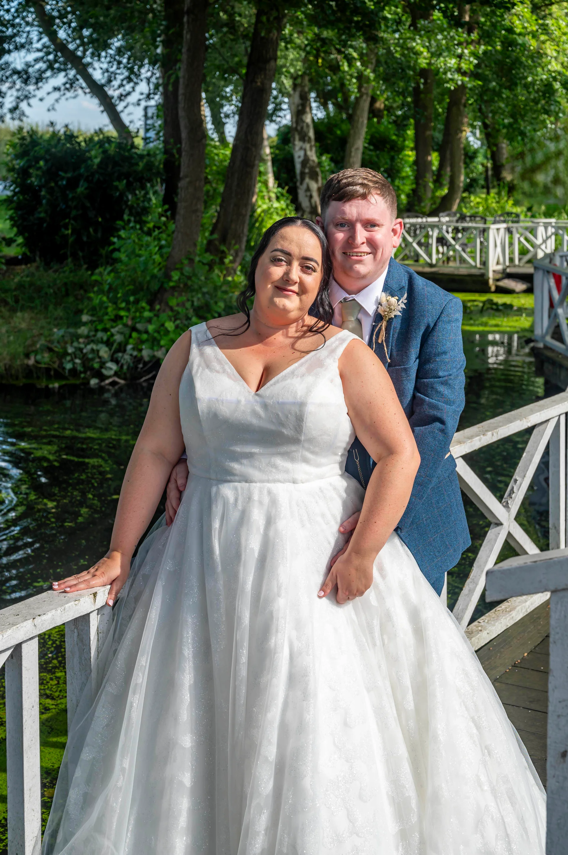 A newlywed couple poses lovingly on a white wooden bridge over a river, surrounded by lush green trees and foliage in the summer sunshine.