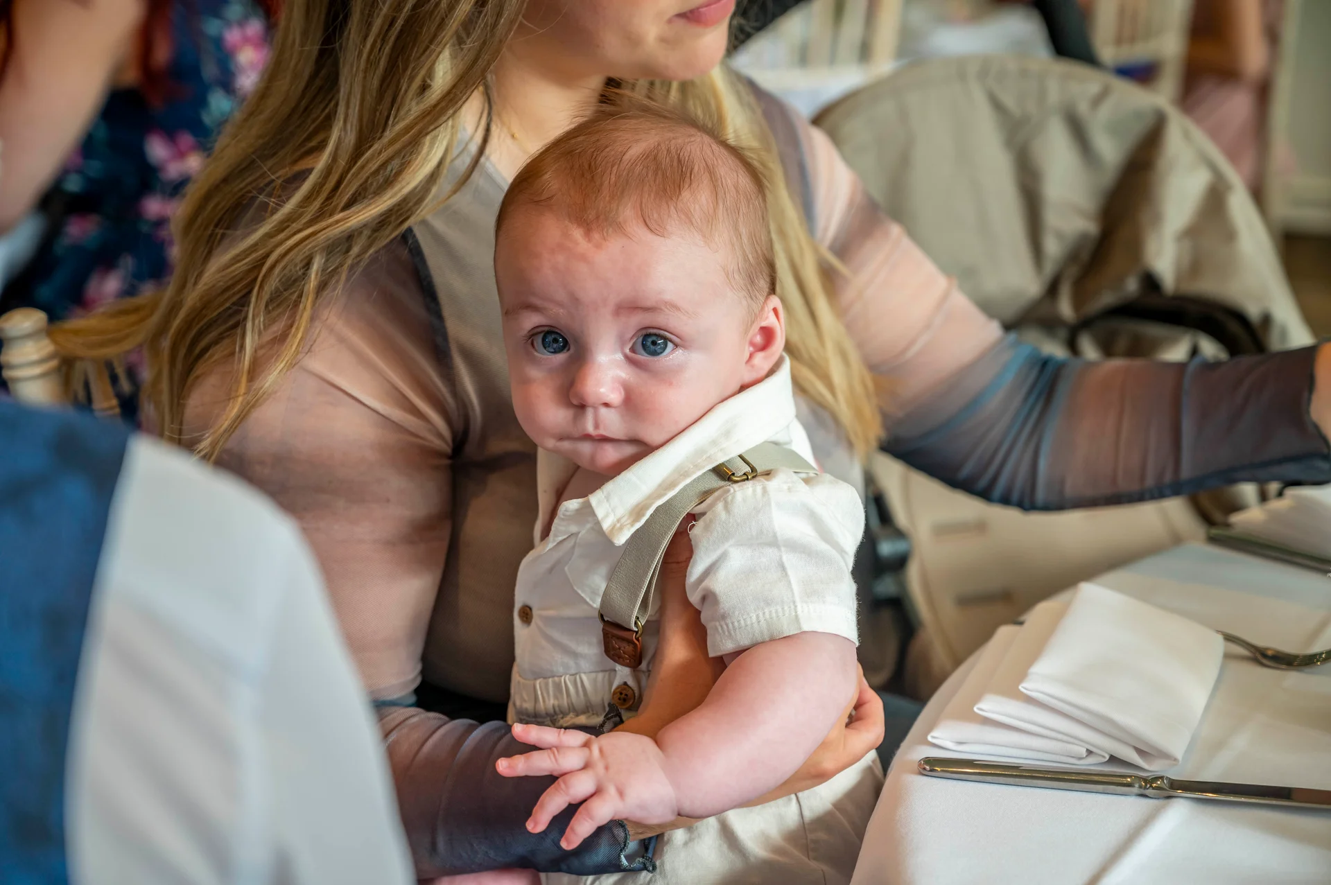 A cute baby wearing a little shirt and braces is held by a guest at a wedding reception table.