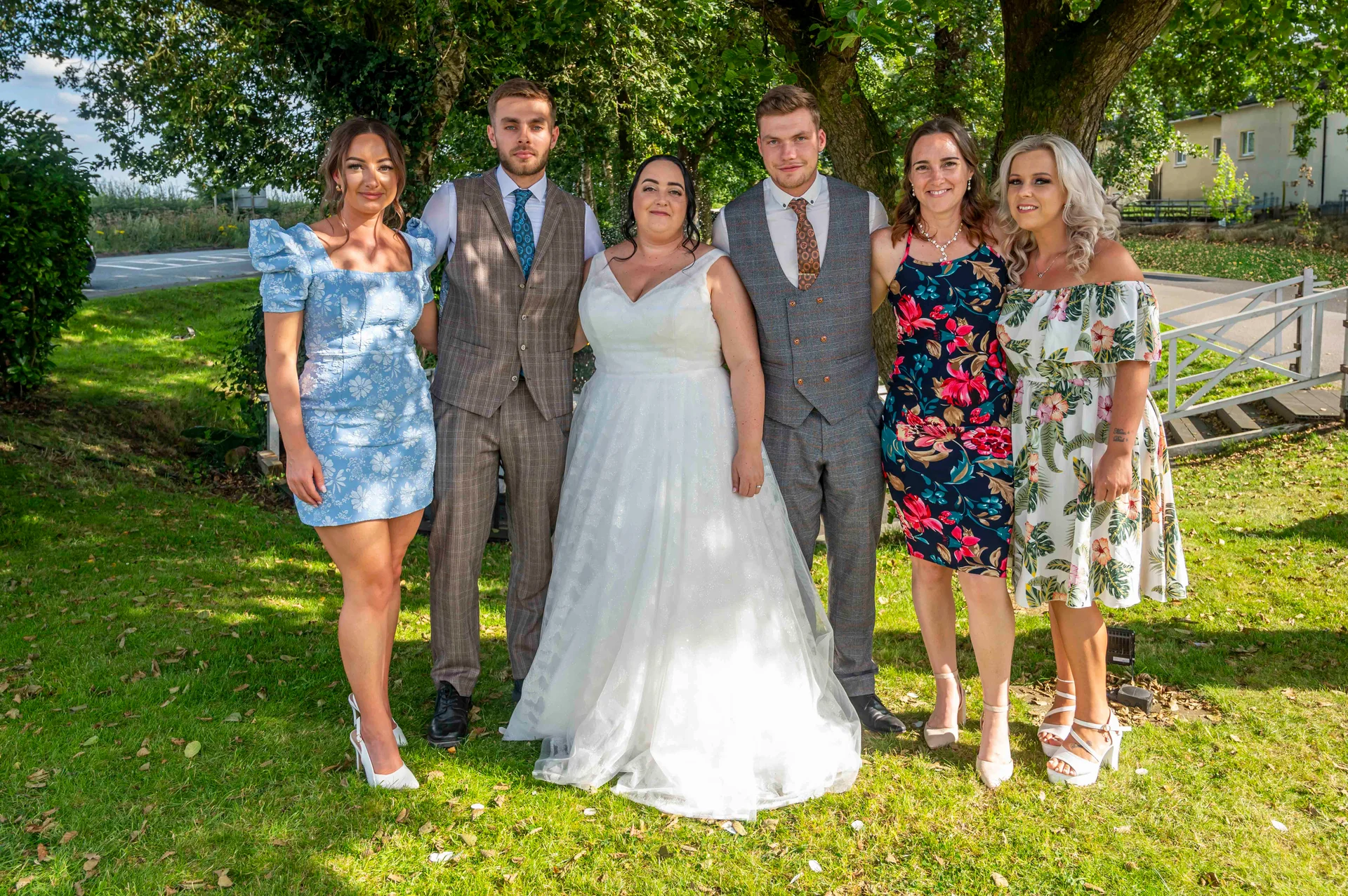 A bridal party and guests pose together on a sunlit lawn outdoors during a summer wedding celebration.