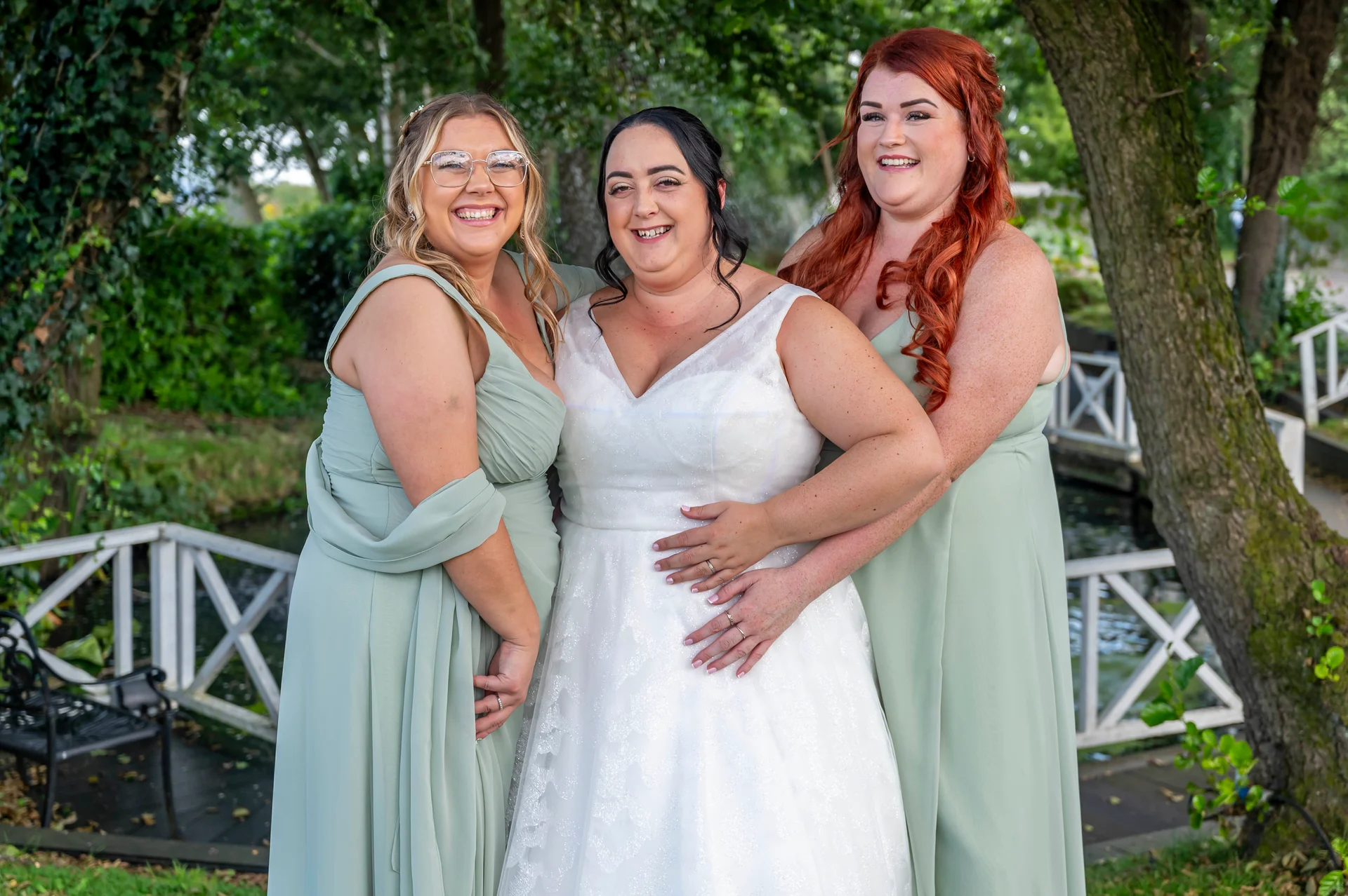 A smiling bride stands outdoors flanked by two bridesmaids wearing sage green dresses amidst lush garden foliage.