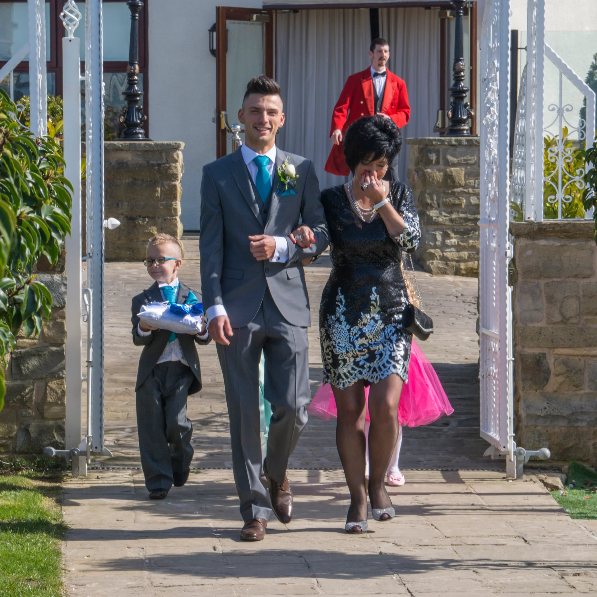 A smiling groom in a grey suit walks arm in arm with the mother of the bride outdoors on a sunny day, accompanied by a young page boy carrying a ring pillow.
