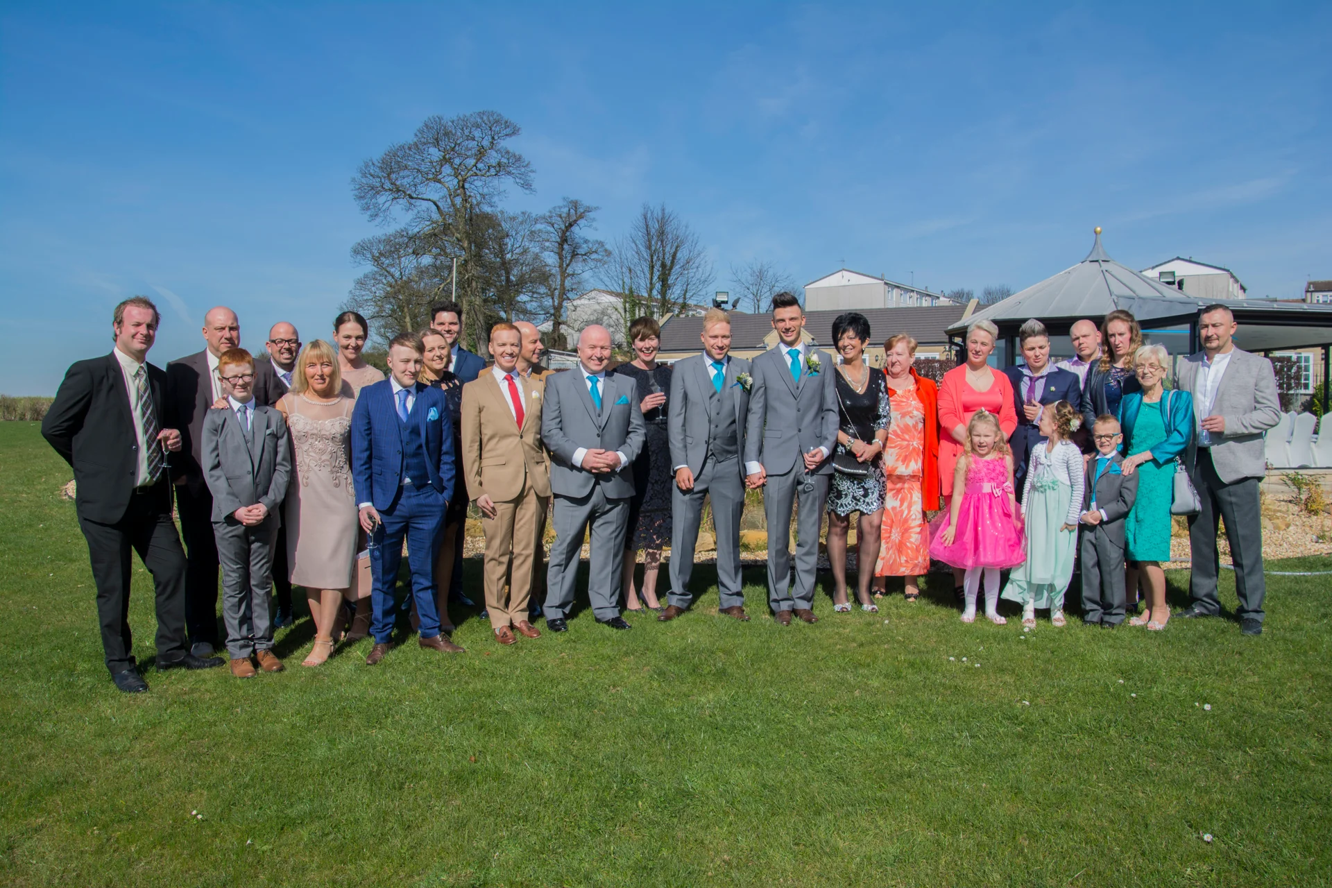 A large wedding party and family group pose smilingly outdoors on a green lawn under a clear blue sky on a sunny day.