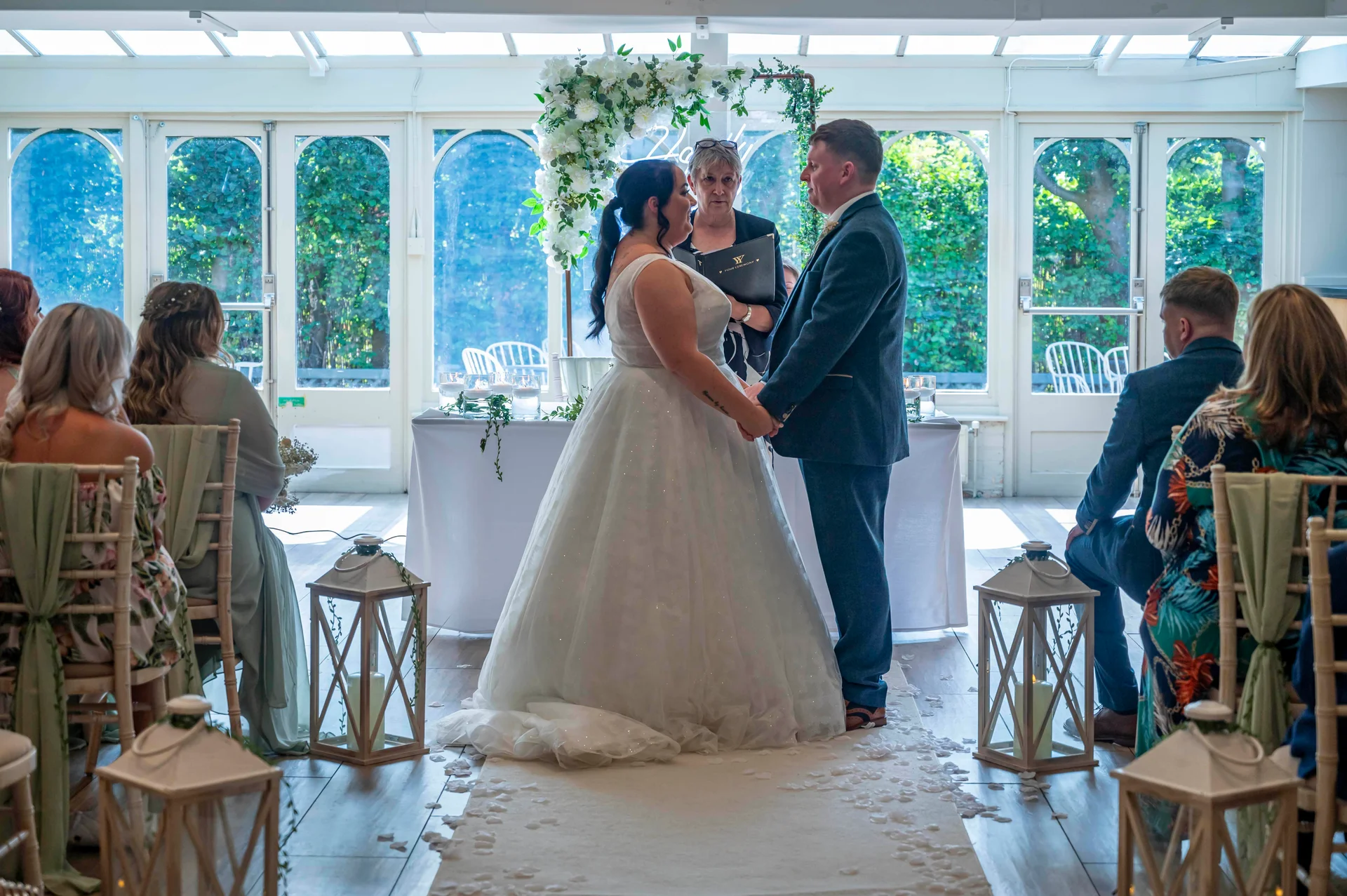 A bride and groom exchange vows during an indoor civil ceremony inside a bright orangery venue decorated with a floral arch and lanterns lining the aisle.