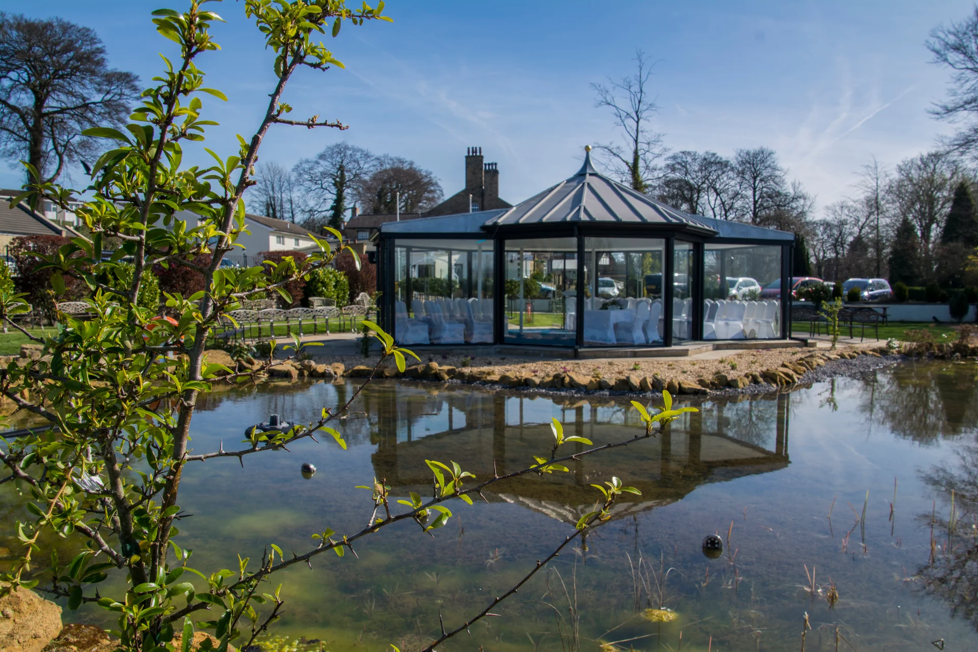 An elegant glass orangery sits by a tranquil ornamental lake under a bright blue spring sky, reflecting perfectly in the calm water.