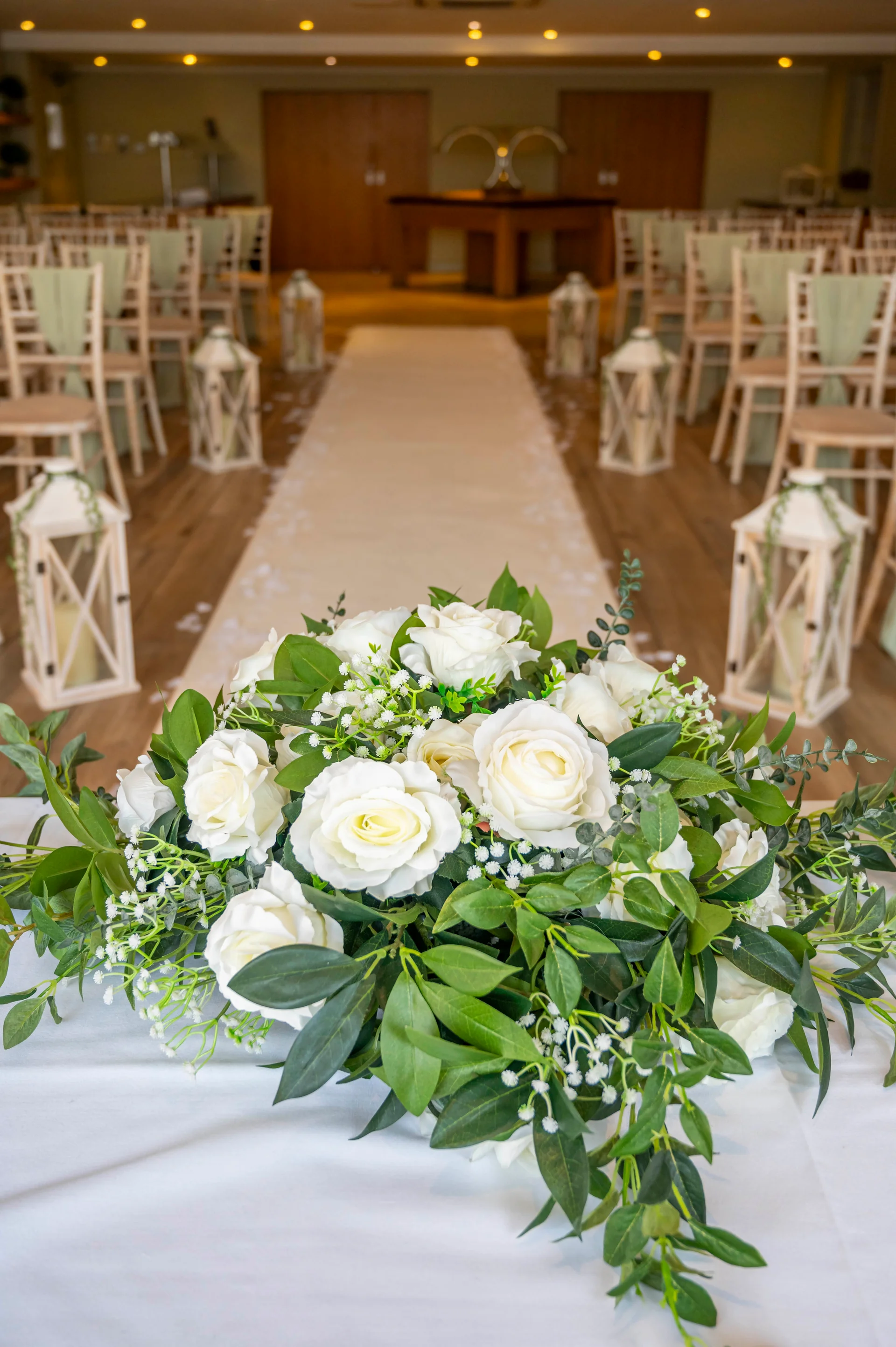 An indoor wedding ceremony venue features a central aisle lined with wooden lanterns and a lush white rose and greenery floral arrangement in the foreground.