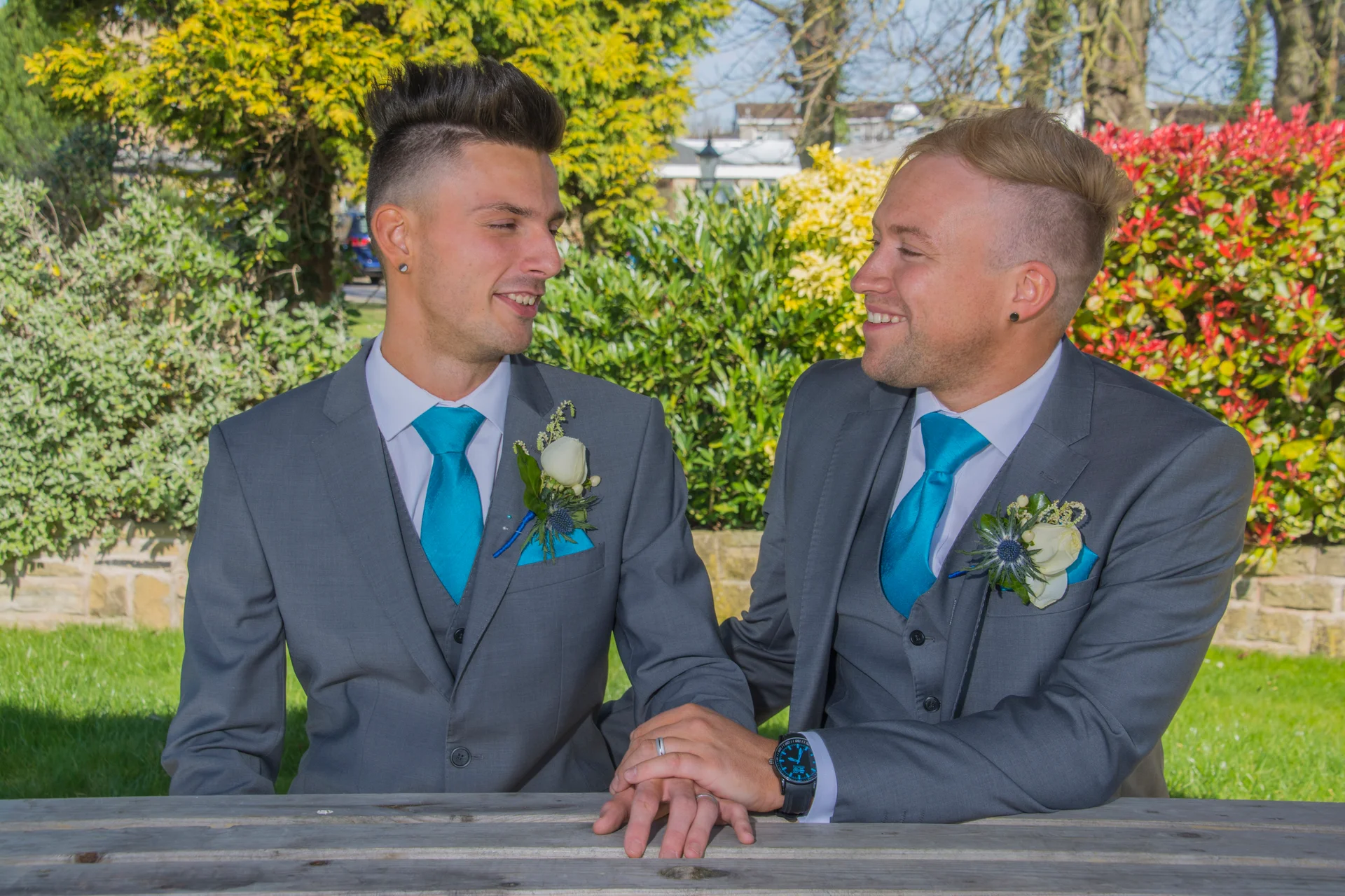 Two smiling grooms in matching grey three-piece suits and vibrant turquoise ties sit outdoors together, holding hands and looking lovingly at each other.